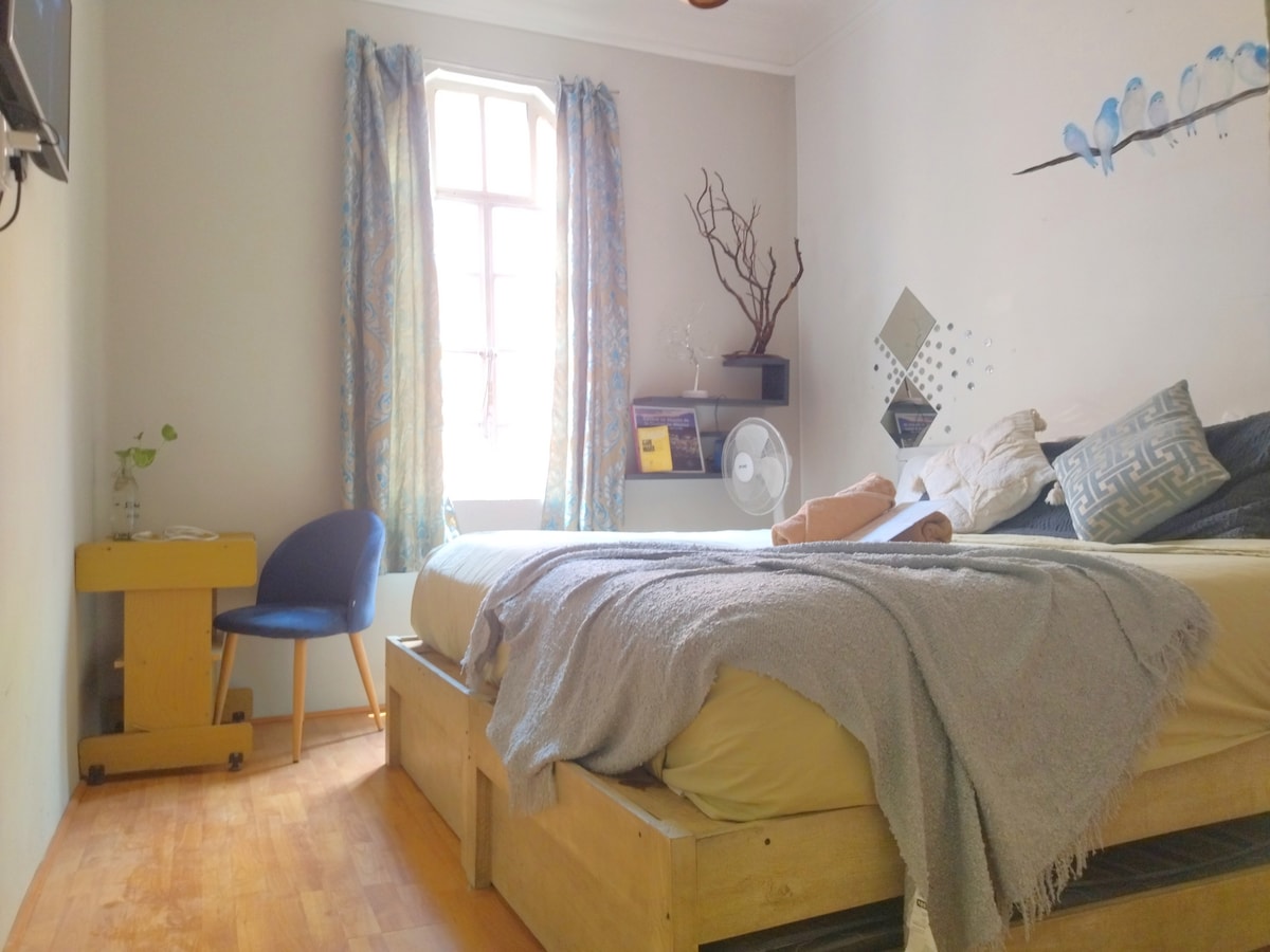 A private room features a king-size bed with a cozy blanket and decorative pillows. A small chair and table are positioned next to the bed. Natural light enters through a window, complemented by patterned curtains. A stand fan is visible, along with minimal decorative elements.