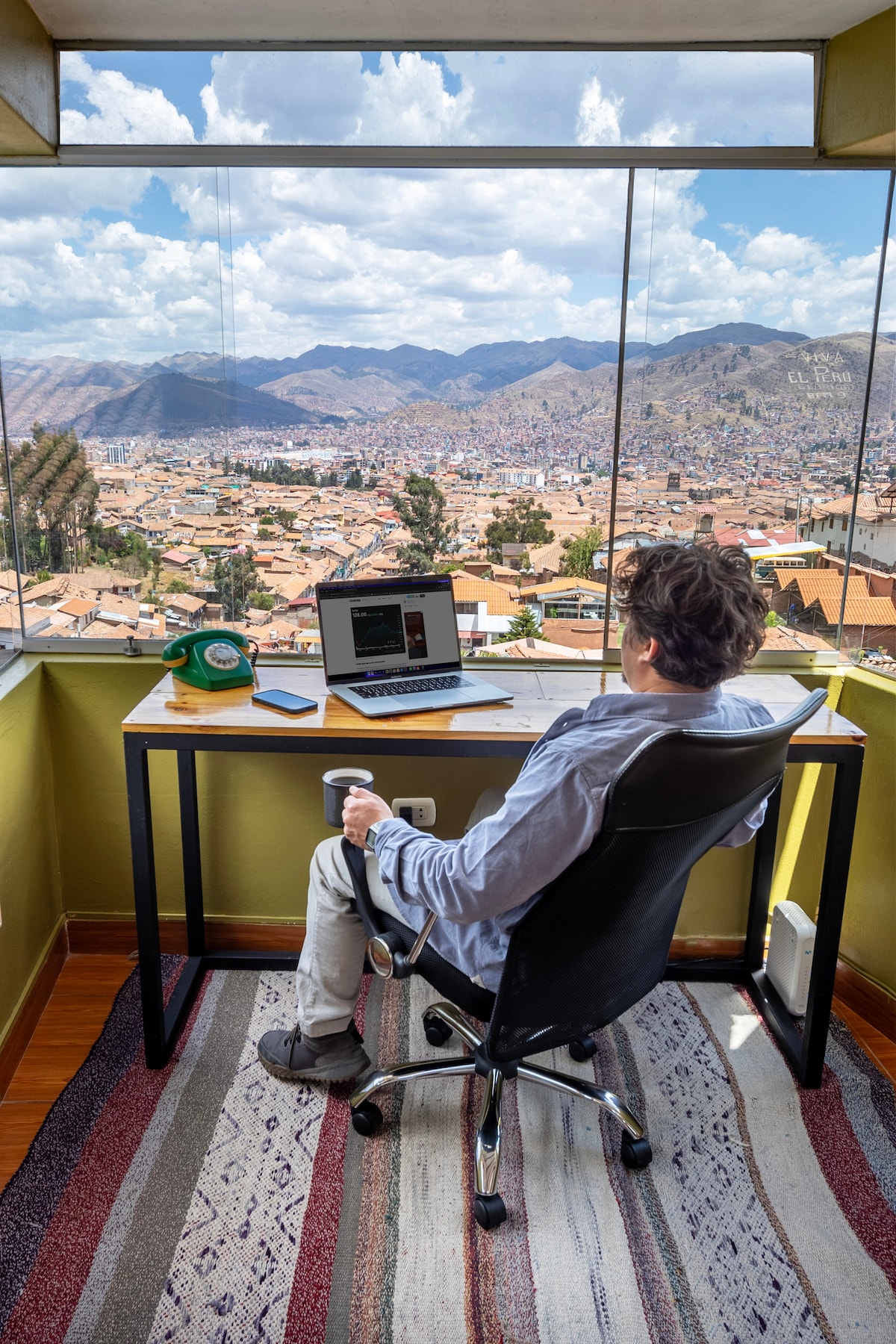 A workspace is visible with a desk positioned by large windows showcasing a panoramic view of Cusco and the surrounding mountains. A person sits comfortably in an ergonomic chair, facing a laptop and a phone placed on the desk, while colorful rugs adorn the floor.