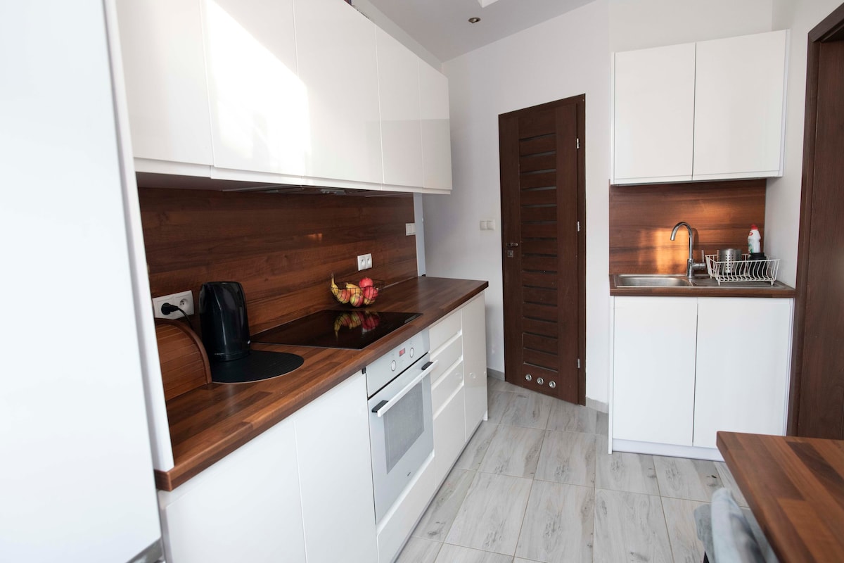 A modern kitchen features sleek white cabinetry paired with warm wood accents. Essential appliances include an oven and a stovetop, while a practical sink is situated against the wall. A small dining table provides space for three, complemented by a decorative bowl of fruit.