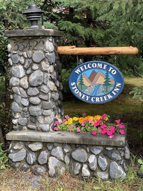 A stone monument features the welcoming sign of Stoney Creek Inn, adorned with a colorful flower bed. The vibrant flowers in shades of pink and yellow contrast with the natural stonework and greenery surrounding the area, providing a friendly entrance to the Inn.