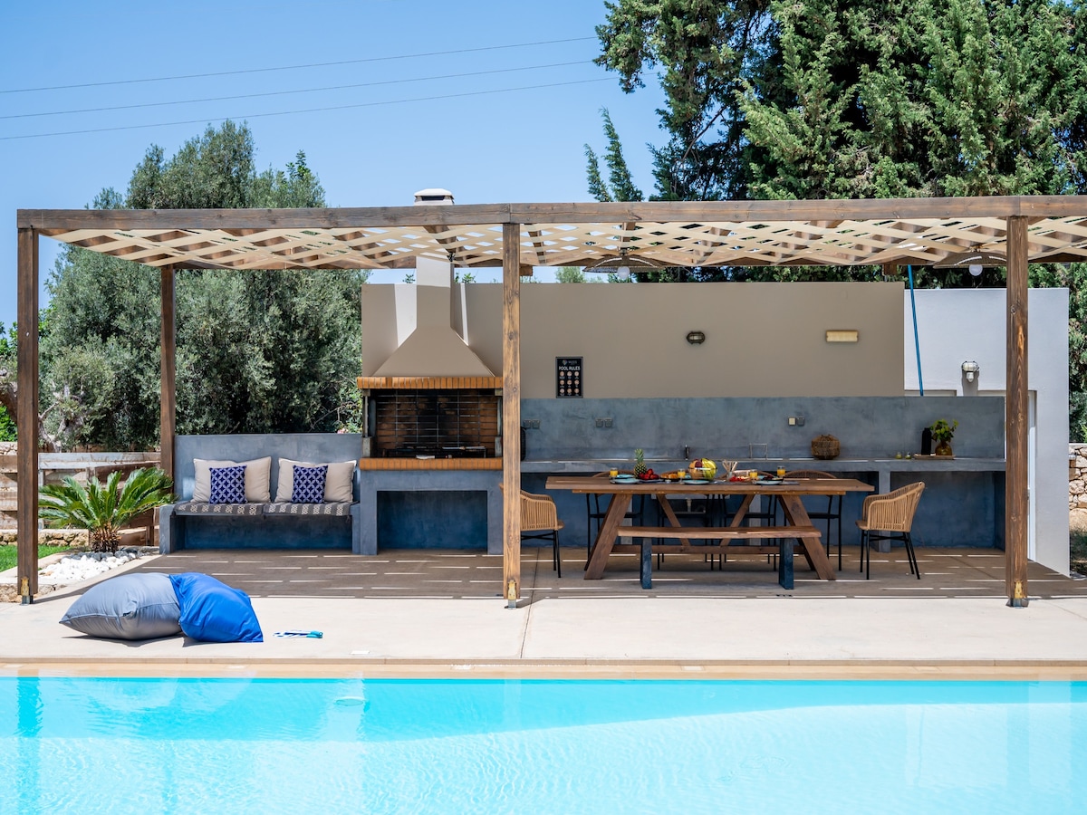 An outdoor dining area features a large table with wooden chairs, set beneath a pergola for shade. A built-in barbecue grill is visible at one side, complemented by a cushioned seating area. The swimming pool reflects the clear sky, enhancing the inviting atmosphere.
