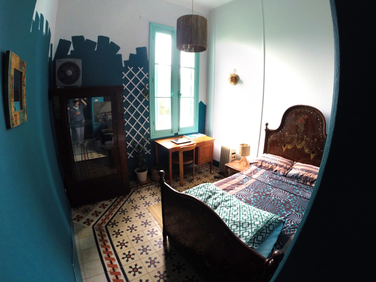 A cozy room features an ornate bed with patterned bedding, complemented by a vintage wardrobe and a wooden desk. Large windows allow natural light to illuminate the space, revealing intricate tile flooring and artistic wall designs.