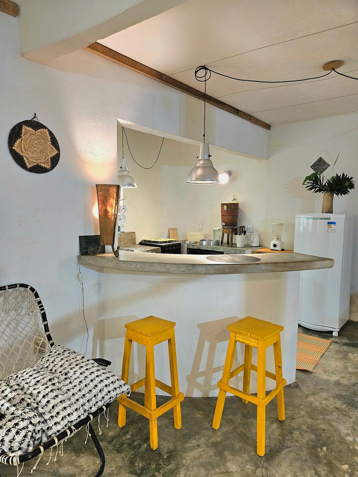 The image shows a compact kitchen with a rounded countertop and two yellow stools. Pendant lights hang from the ceiling, illuminating the space. A white refrigerator is visible in the background, and local artifacts are displayed on the walls, adding character to the area.