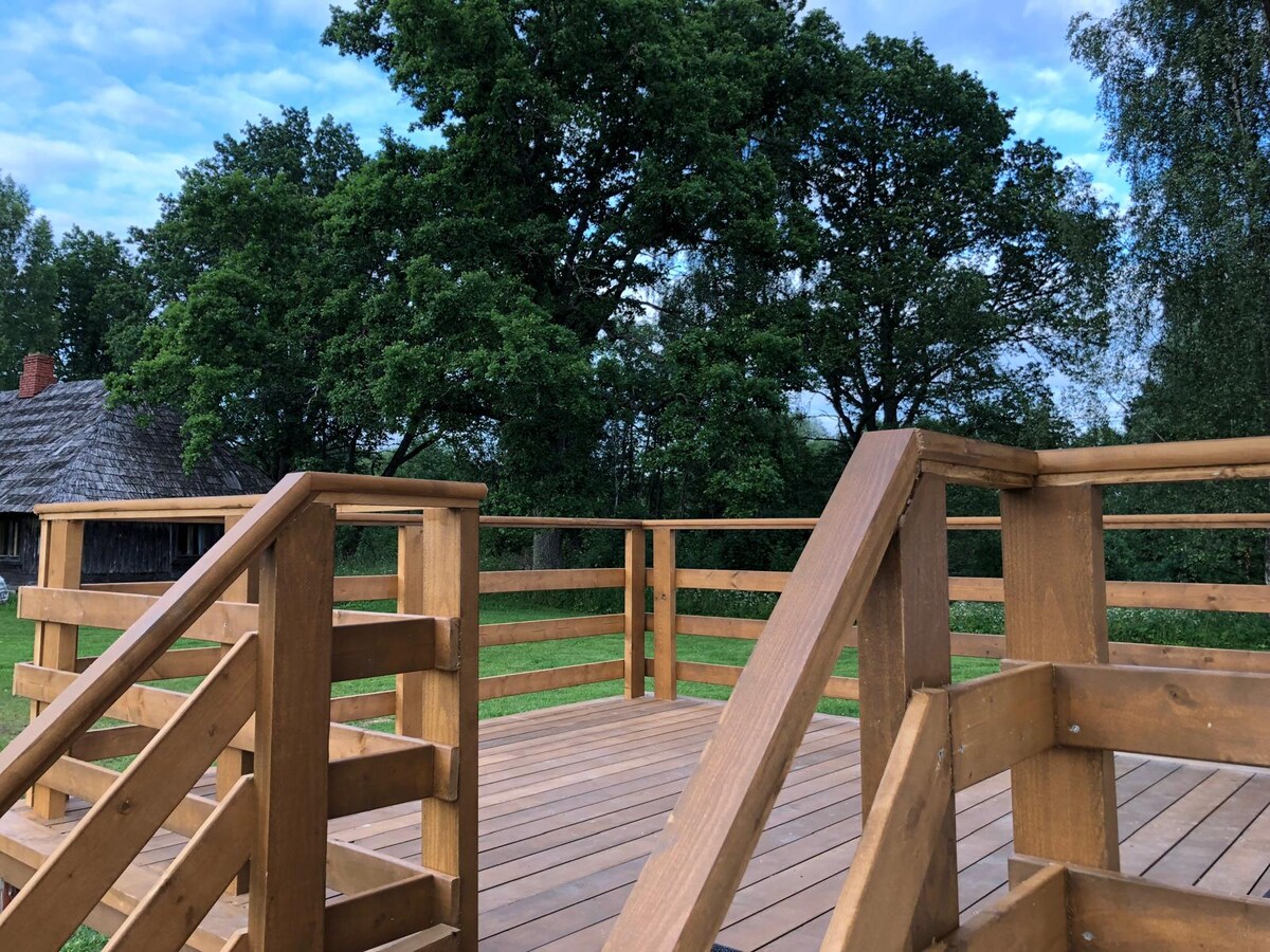 A wooden deck is prominently featured, surrounded by lush greenery and trees. Steps lead down from the deck, and the smooth, natural wood grain is visible. The open space invites relaxation, while the scenery adds a touch of nature's beauty.