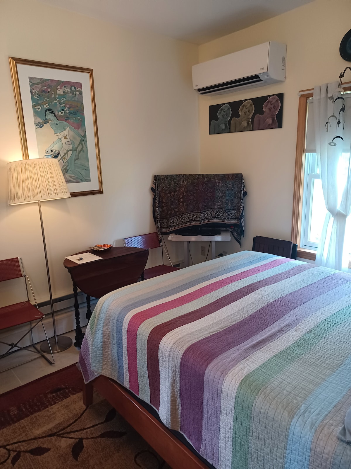A cozy studio bedroom features a spacious bed draped with a striped quilt, complemented by a decorative rug. Natural light enters through a window adorned with soft curtains. An art piece hangs above the bed, with a small table and two chairs accommodated nearby.
