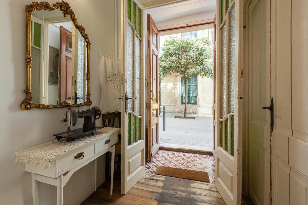 An inviting entryway features vintage elements, including a decorative mirror and a sewing machine on a white table. Doors open to a view of a courtyard tree, while patterned tiles and wooden floors add character to the space.