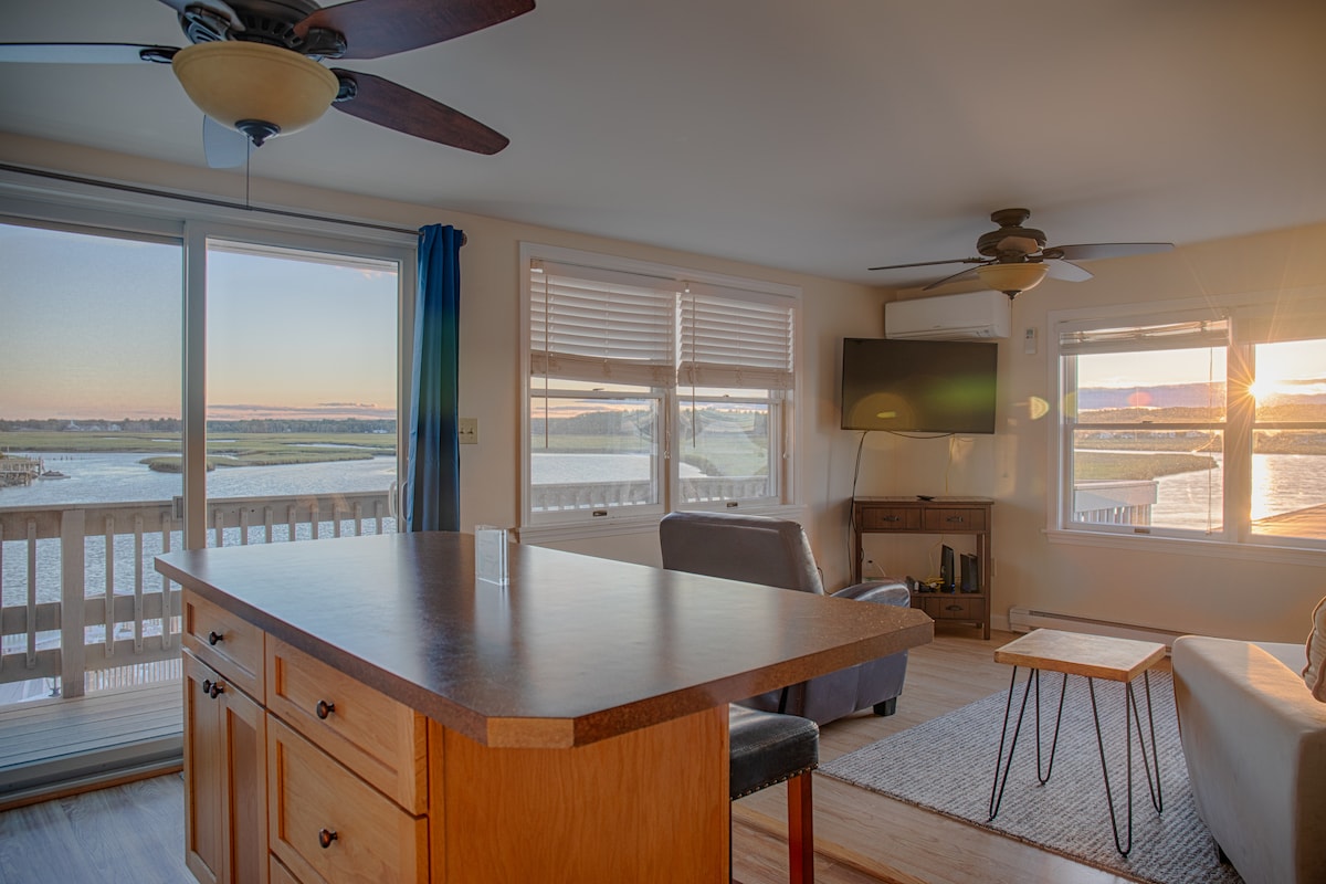The interior space features a kitchen island with wooden cabinetry, adjacent to a living area with a gray sofa and a small wooden side table. Large windows offer expansive views of the Rachel Carson Preserve, with sunlight reflecting off the water.