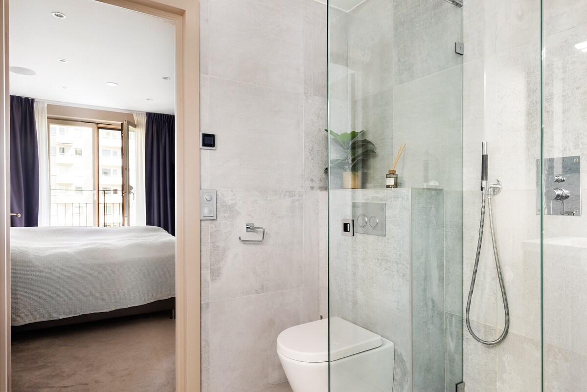 A modern bathroom is shown, featuring a glass shower enclosure and a toilet. Soft lighting complements the neutral-toned walls and tiled flooring. A glimpse of the bedroom is visible beyond, showcasing a spacious bed and large windows dressed with dark curtains.