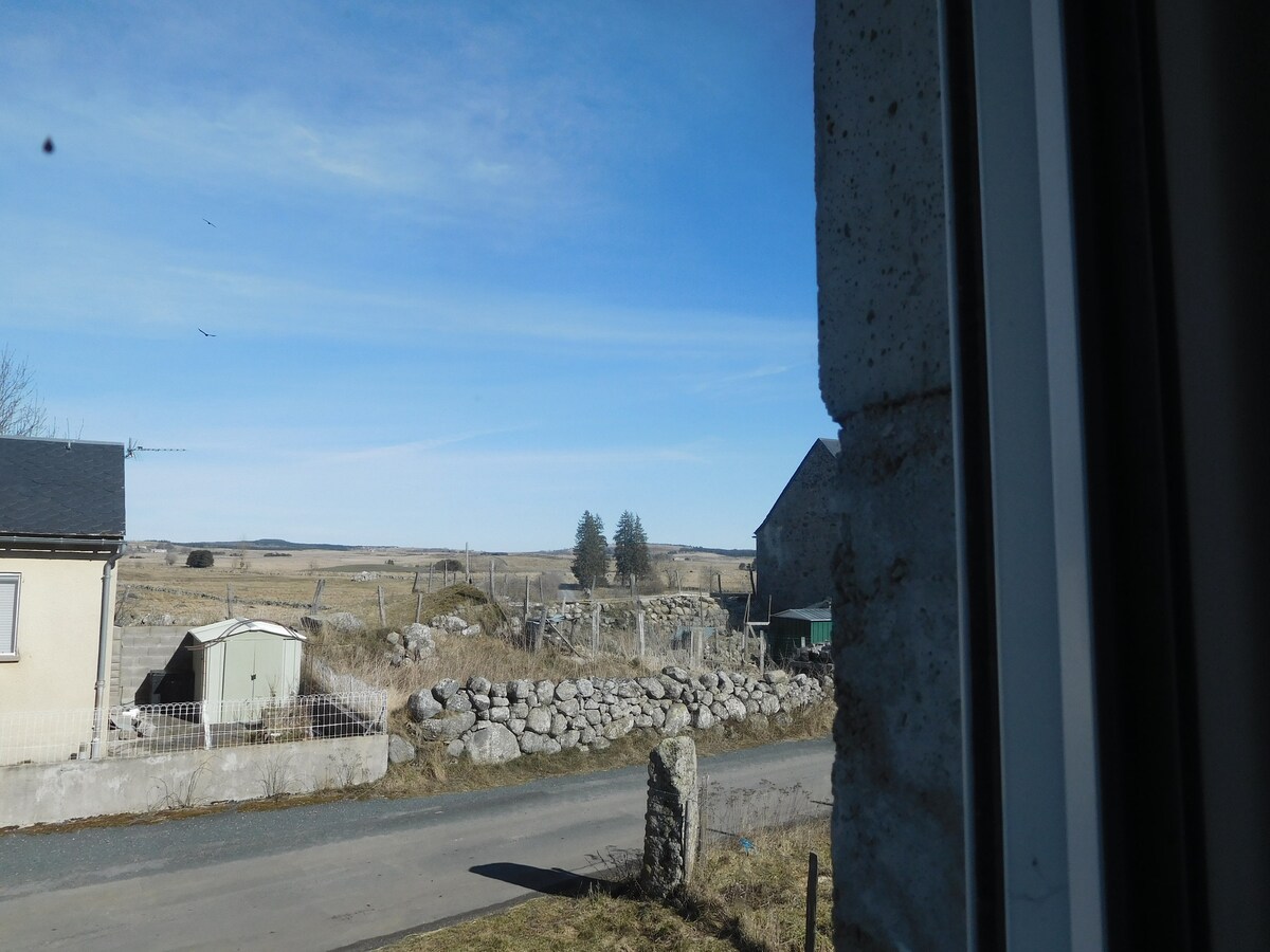A view from a window showcases an open rural landscape with rolling hills and a clear blue sky. A dirt road runs parallel to a stone wall, while a small shed and distant structures are visible among the fields.