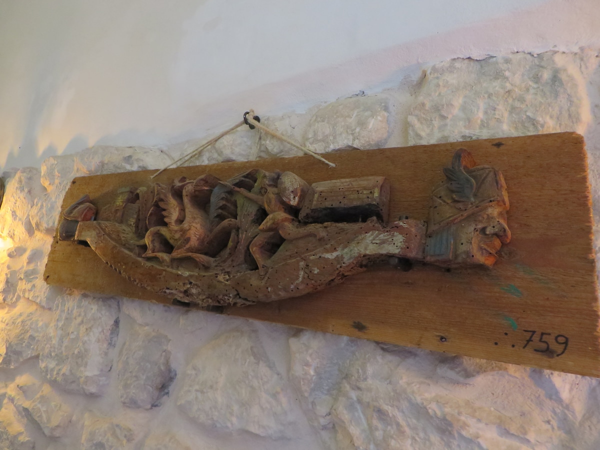 A wooden wall carving is displayed prominently against a stone wall. The artwork features intricate depictions of animals and nature, showcasing detailed craftsmanship. Soft lighting accentuates the textures and colors of the carving, creating a focal point in the room.