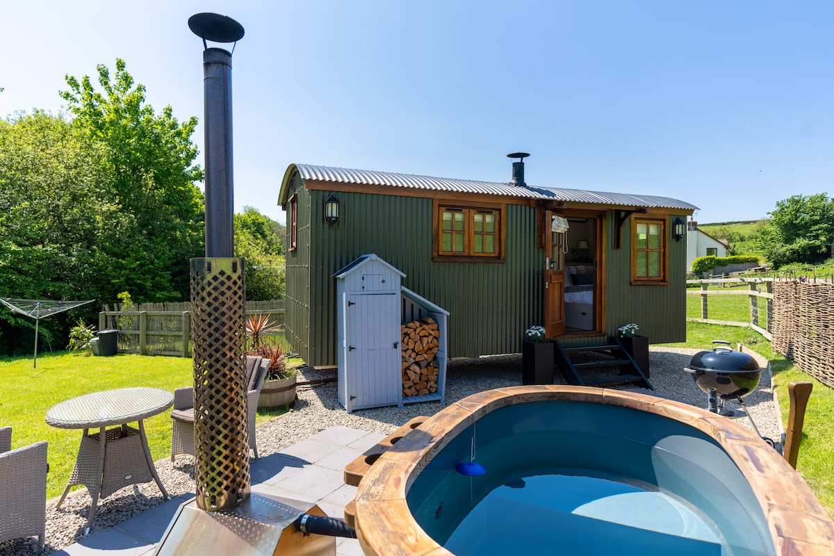 A charming Shepherd's Hut is set against a clear blue sky, showcasing an outdoor wood-burning hot tub framed by a wooden deck. Comfortable patio furniture is arranged nearby, and a small wooden shed displays firewood. Lush greenery borders the scene, enhancing the rural ambience.