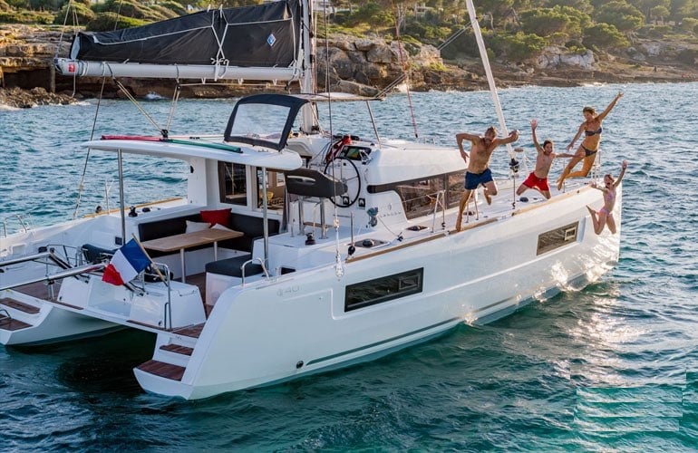 A modern catamaran is anchored in clear turquoise waters, surrounded by scenic coastal landscapes. Four guests are seen joyfully jumping off the side of the boat, emphasizing a lively atmosphere. The boat features a spacious deck with seating areas, while the sails are raised against the backdrop of a blue sky.