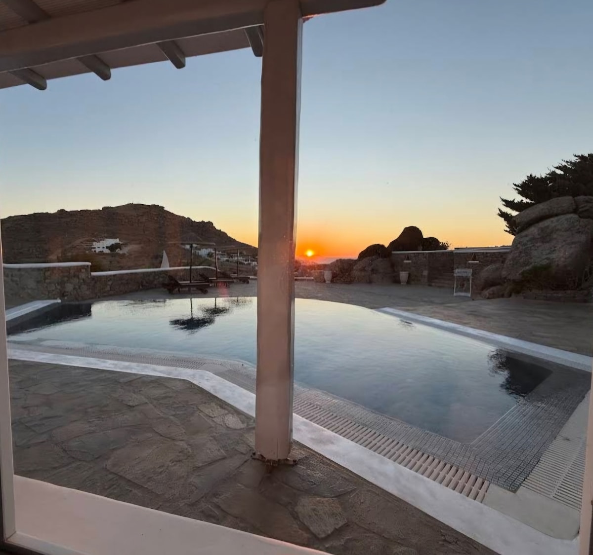 A serene outdoor pool area is captured at sunset, showcasing gentle ripples on the water's surface. The scene is framed by a stone wall and the silhouette of surrounding hills. Light from the setting sun casts a warm glow, enhancing the tranquil atmosphere.