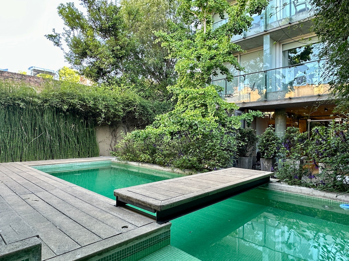 A serene outdoor space features a calm swimming pool, bordered by lush greenery. A wooden deck extends over the water, providing a relaxing area for sunbathing or socializing. The building is visible in the background, showcasing modern architectural elements.