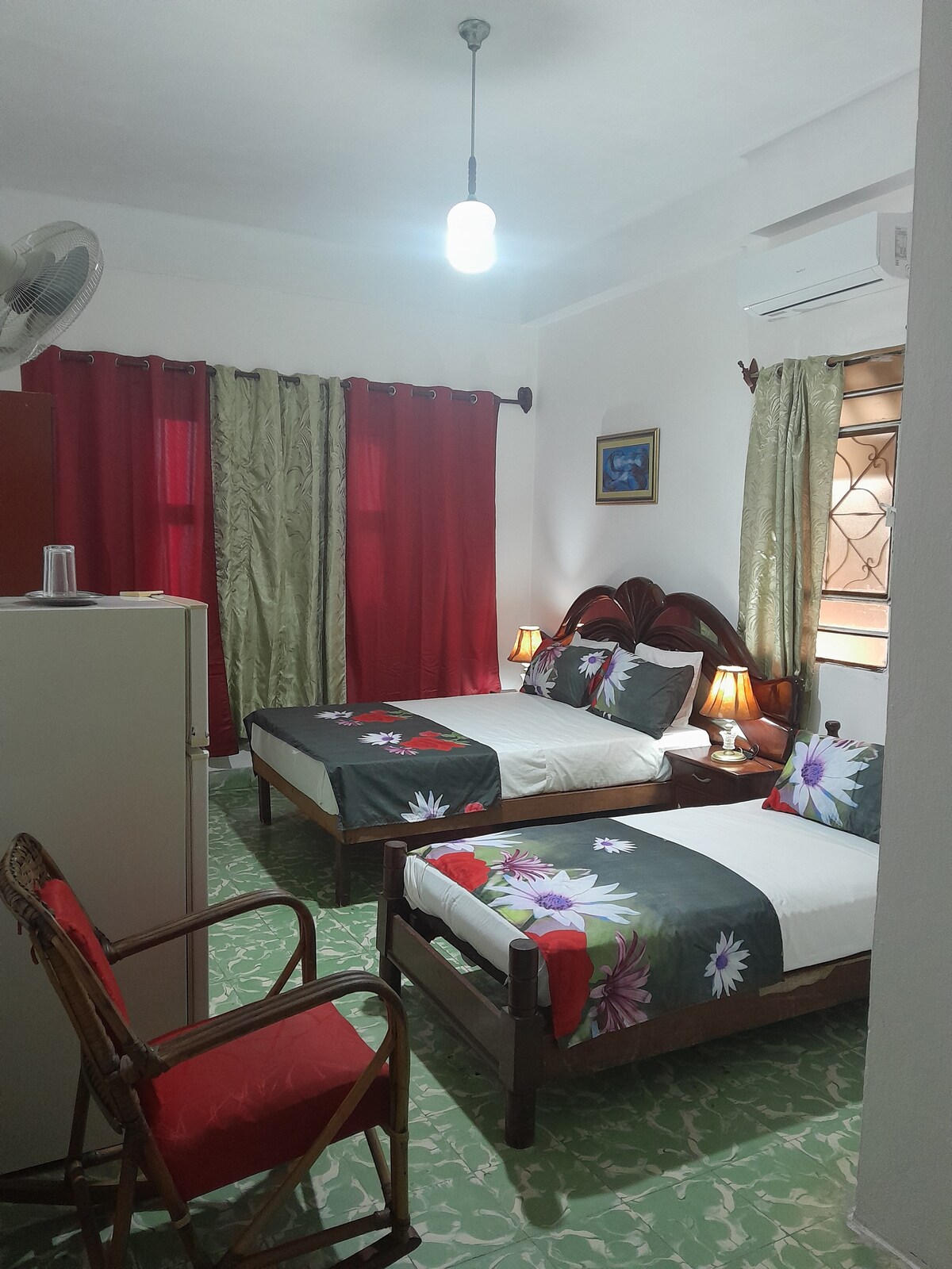 The room features two beds with floral-patterned bedspreads, providing a bright and welcoming atmosphere. A wardrobe stands against one wall, and a comfortable chair is positioned nearby. Natural light filters through the windows, complemented by elegant curtains and lamps on each bedside table.