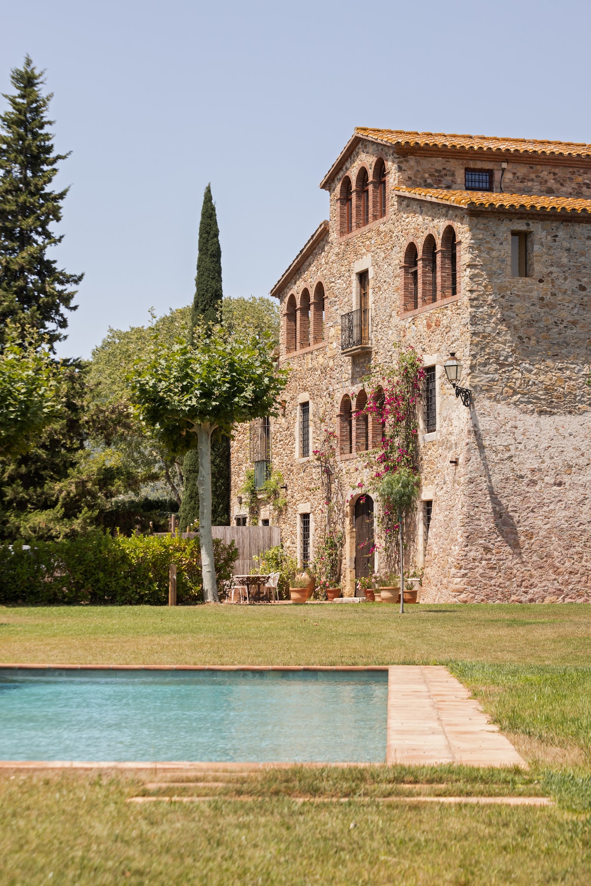 A historic stone farmhouse with distinctive architectural features is set amidst a lush green landscape. The inviting salt-water pool reflects the clear sky, while well-maintained gardens adorned with trees and flowering plants enhance the serene setting.