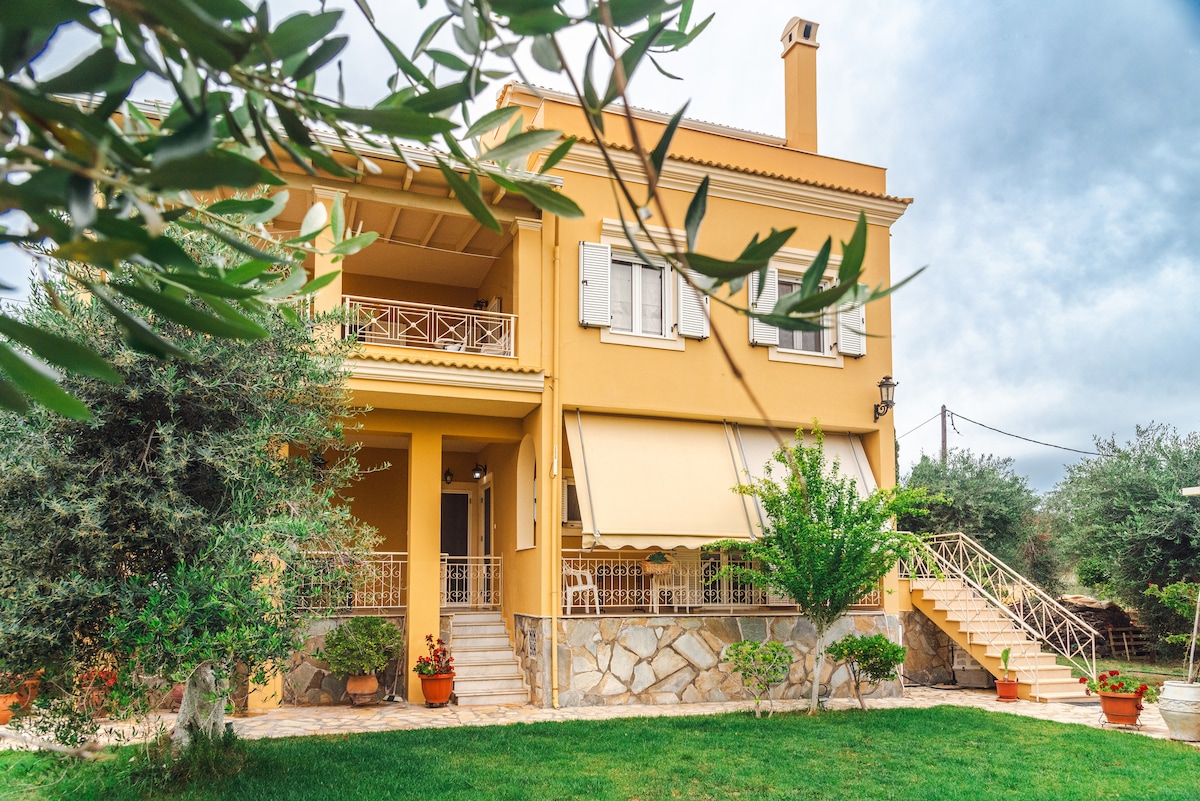 A multi-story yellow house is surrounded by greenery, featuring olive trees and potted plants. A front terrace is accessible via a stone staircase. Large windows and shutters are visible, while a sunshade covers part of the porch, providing a welcoming entrance.