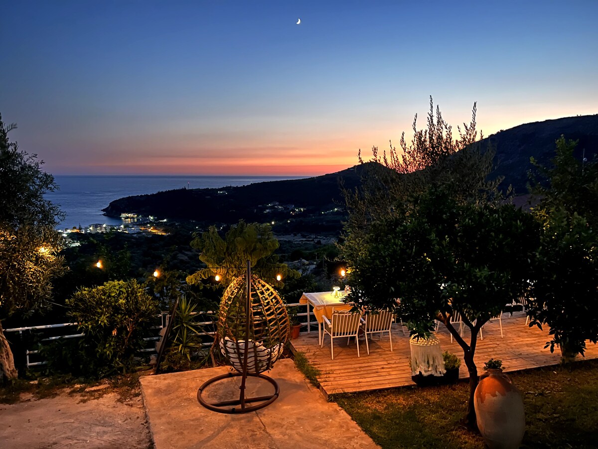 A patio area is visible at sunset, showcasing a woven hanging chair and a dining table surrounded by white chairs. Lush greenery borders the space, and distant lights twinkle along the coastline beneath a sky transitioning from orange to deep blue, with a crescent moon evident.