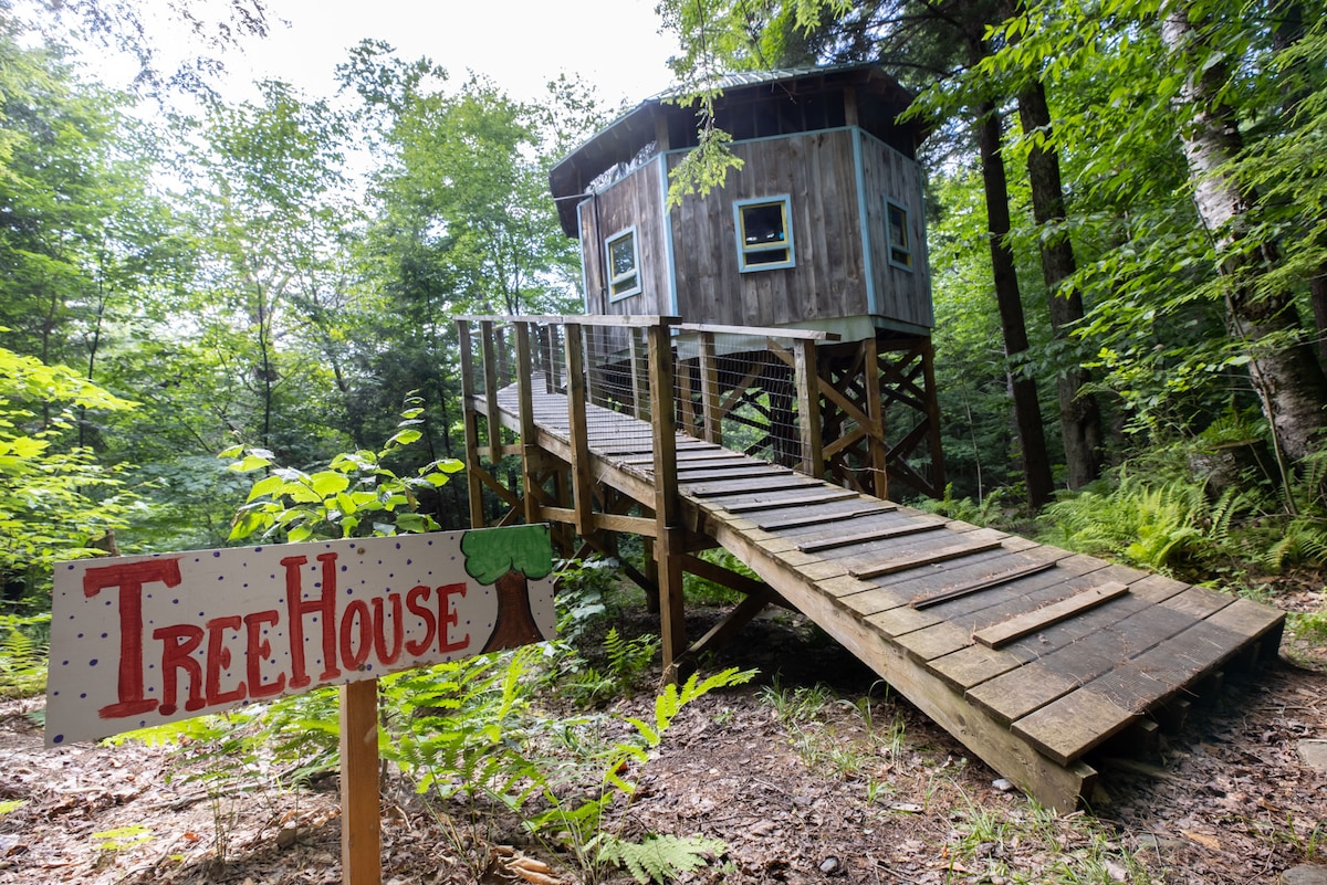 A wooden treehouse is elevated among lush green trees, accessed by a sturdy wooden walkway. A colorful sign labeled 'TreeHouse' stands at the beginning of the path, enhancing the inviting entrance to this nature retreat.