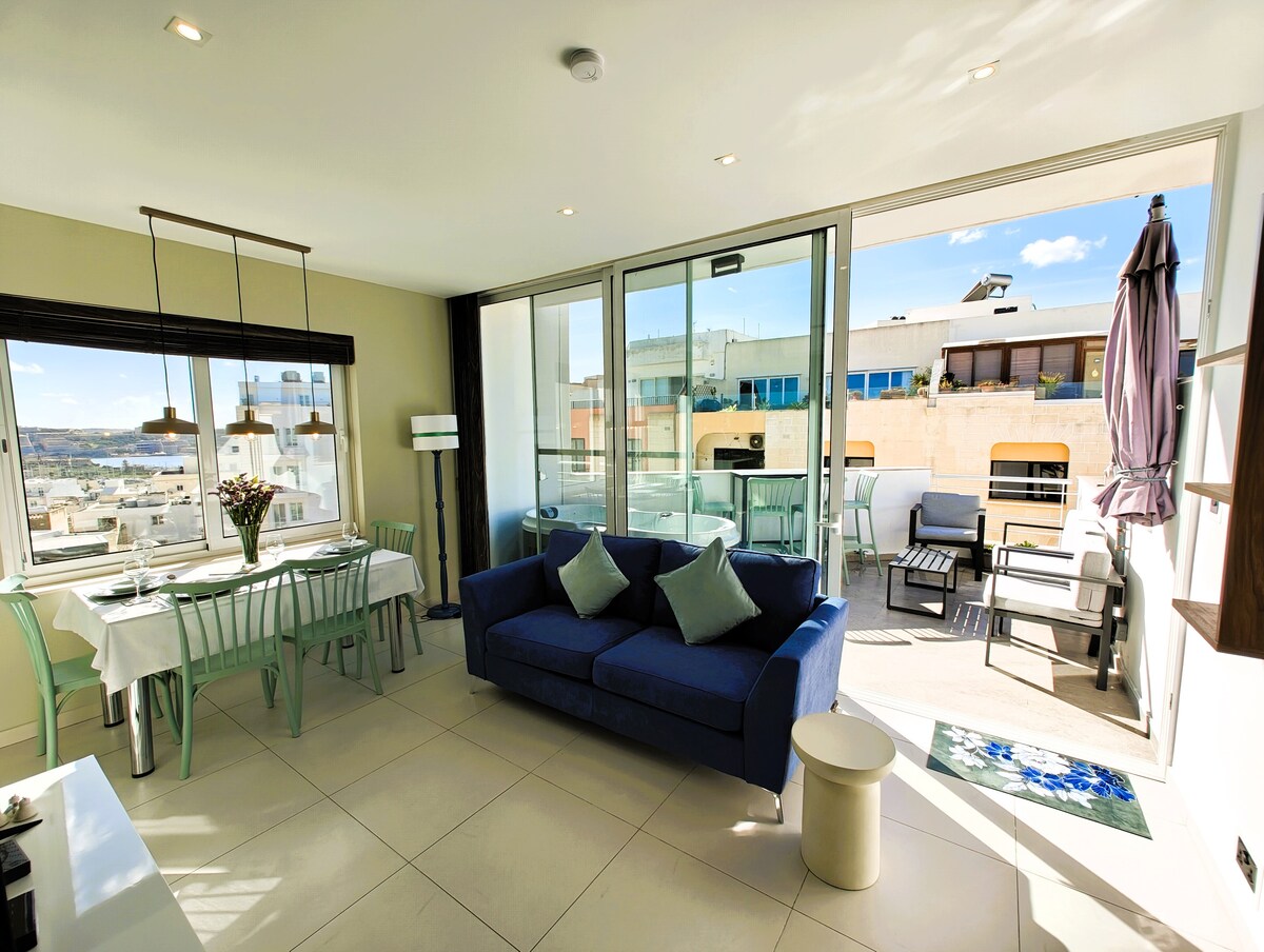 The living and dining area features a light-filled space with large windows offering views of the terrace. A comfortable blue sofa is positioned beside a dining table set for four. The adjoining terrace is furnished with outdoor seating, providing an inviting setting for relaxation.