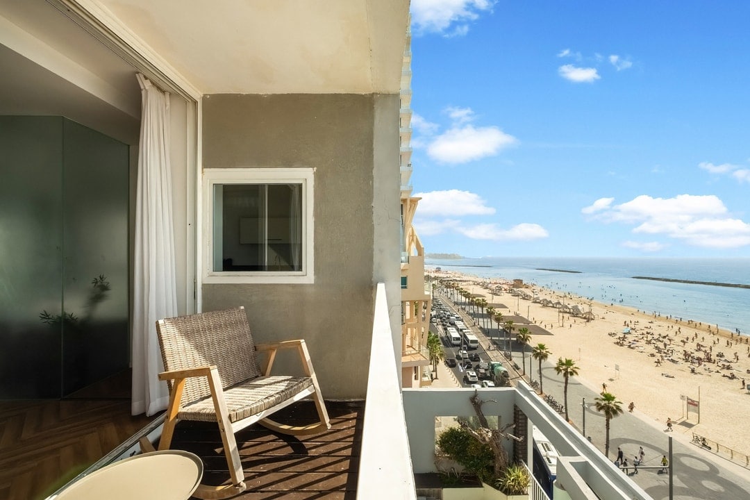 A private balcony features a wooden chair overlooking a sandy beach, with views of the sea and scattered beachgoers. Sunlight brightens the space, and a small table partially visible enhances the outdoor seating area.