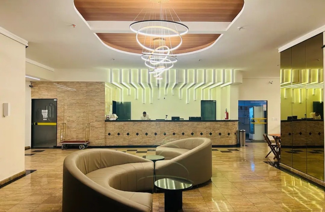 A spacious reception area features elegant circular light fixtures above a sleek curved couch. The warm-toned walls are complemented by modern accents, and a reception desk is visible in the background. Large mirrors enhance the openness of the space.