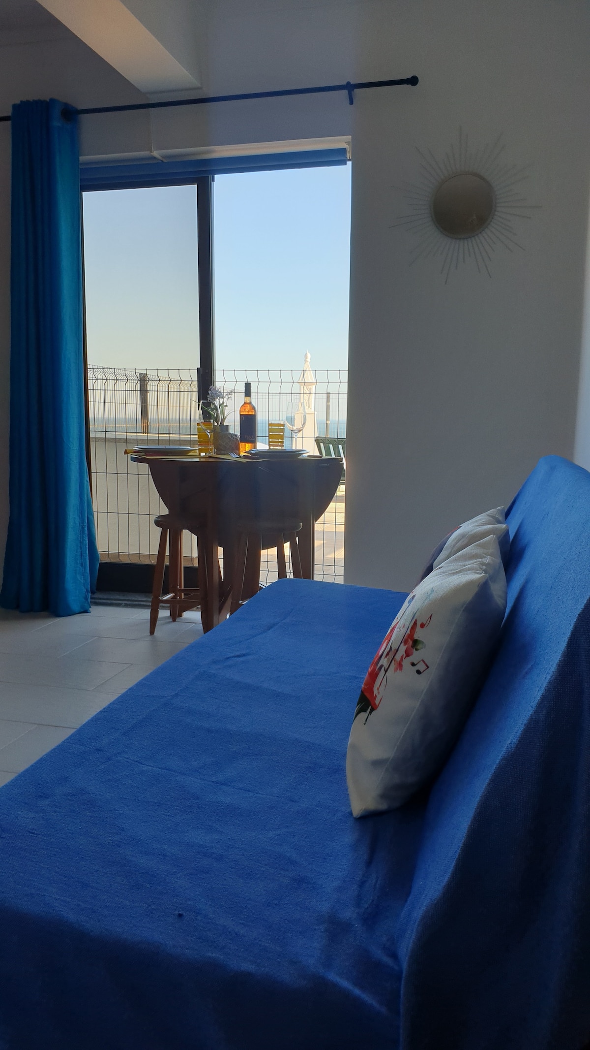 A cozy seating area features a blue couch with a decorative pillow. Natural light fills the space, highlighting a small dining table set for two with a bottle and glasses. A large window offers views of the ocean and outdoor seating.