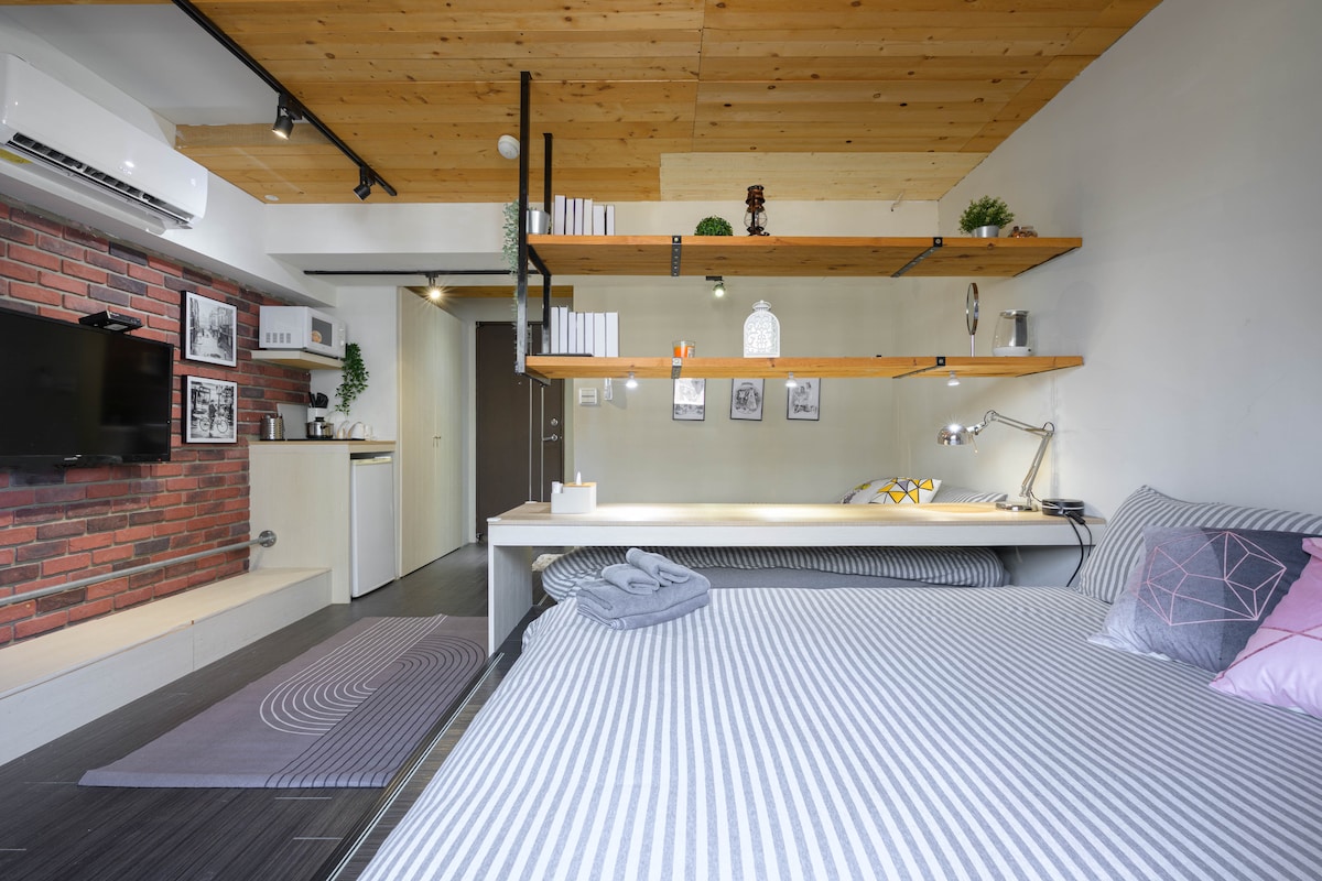 A cozy sleeping area with a spacious bed is complemented by striped bedding and soft towels. Open shelving displays decorative items, while a work desk with a lamp is positioned nearby. A contrasting brick wall adds texture, and a flat-screen TV is mounted for convenience.