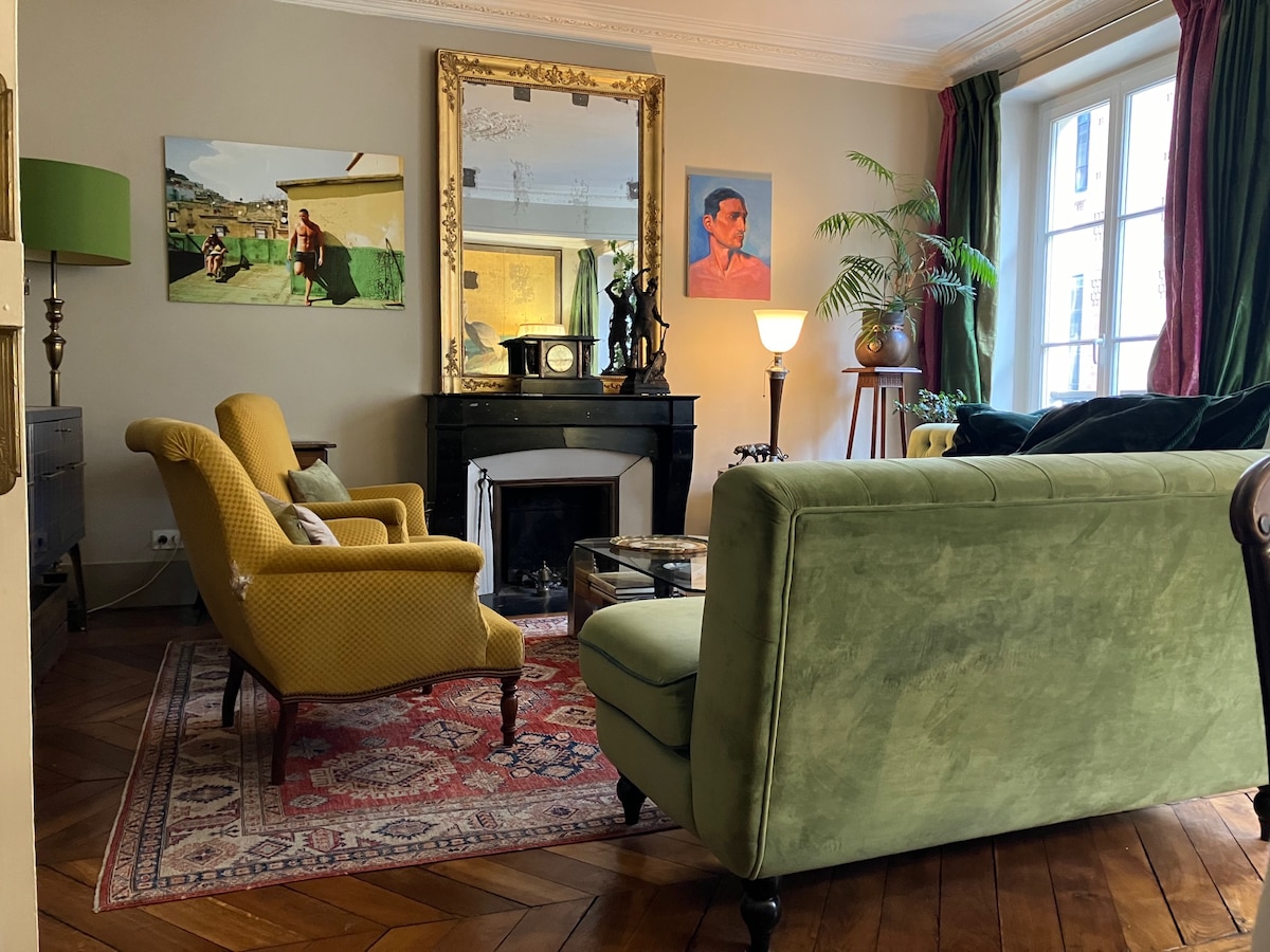A welcoming living room is captured, featuring two yellow armchairs and a green sofa, arranged around a central glass coffee table. A large mirror is mounted above a dark fireplace, and colorful artwork adorns the walls. Natural light filters in through tall windows, enhancing the space.