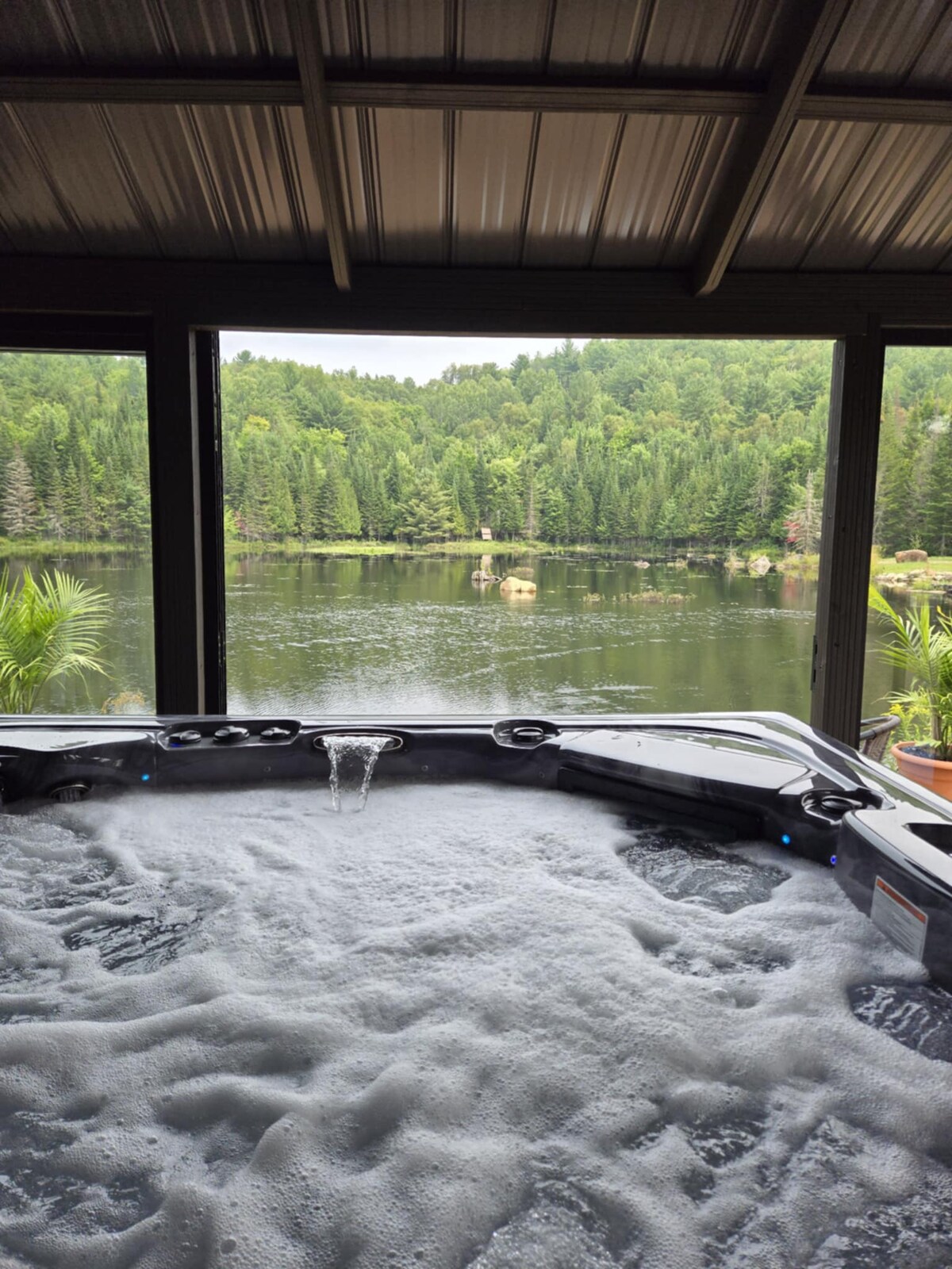 A hot tub filled with bubbling water is positioned in front of large glass windows, providing a view of a serene lake surrounded by lush greenery. The peaceful landscape reflects a tranquil nature setting.