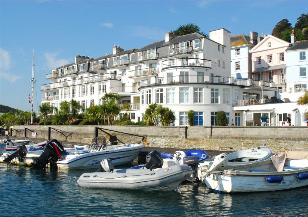 The image showcases a waterfront view of a sleek, modern building surrounded by palm trees. Various boats are moored in the foreground, highlighting the coastal charm. The vibrant blue sky complements the scene, creating a relaxed atmosphere in this popular seaside location.