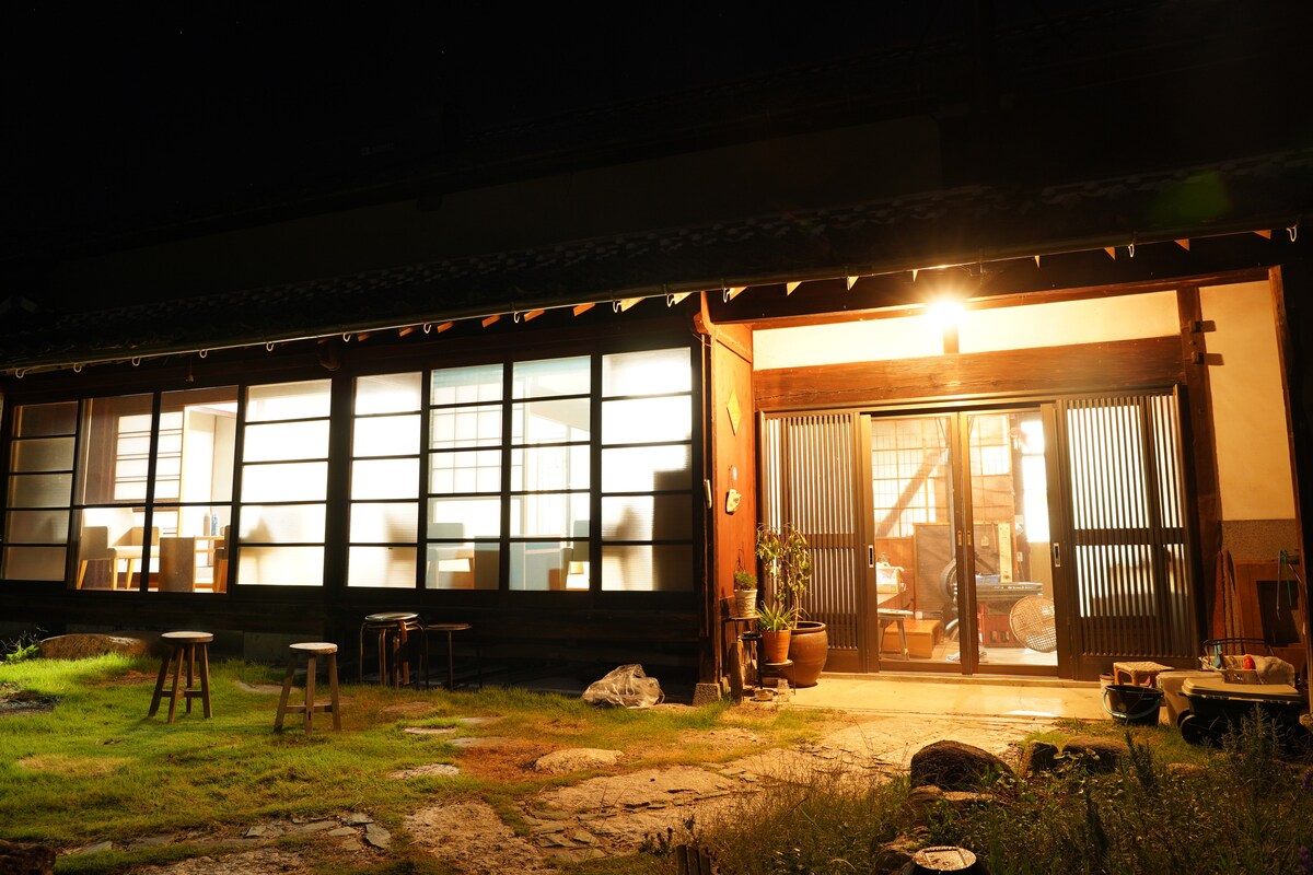 An old traditional house is illuminated at night, showcasing sliding doors that reveal a softly lit interior. Natural grass surrounds the entrance, and small wooden stools are placed outside, adding to the rustic charm of the setting.