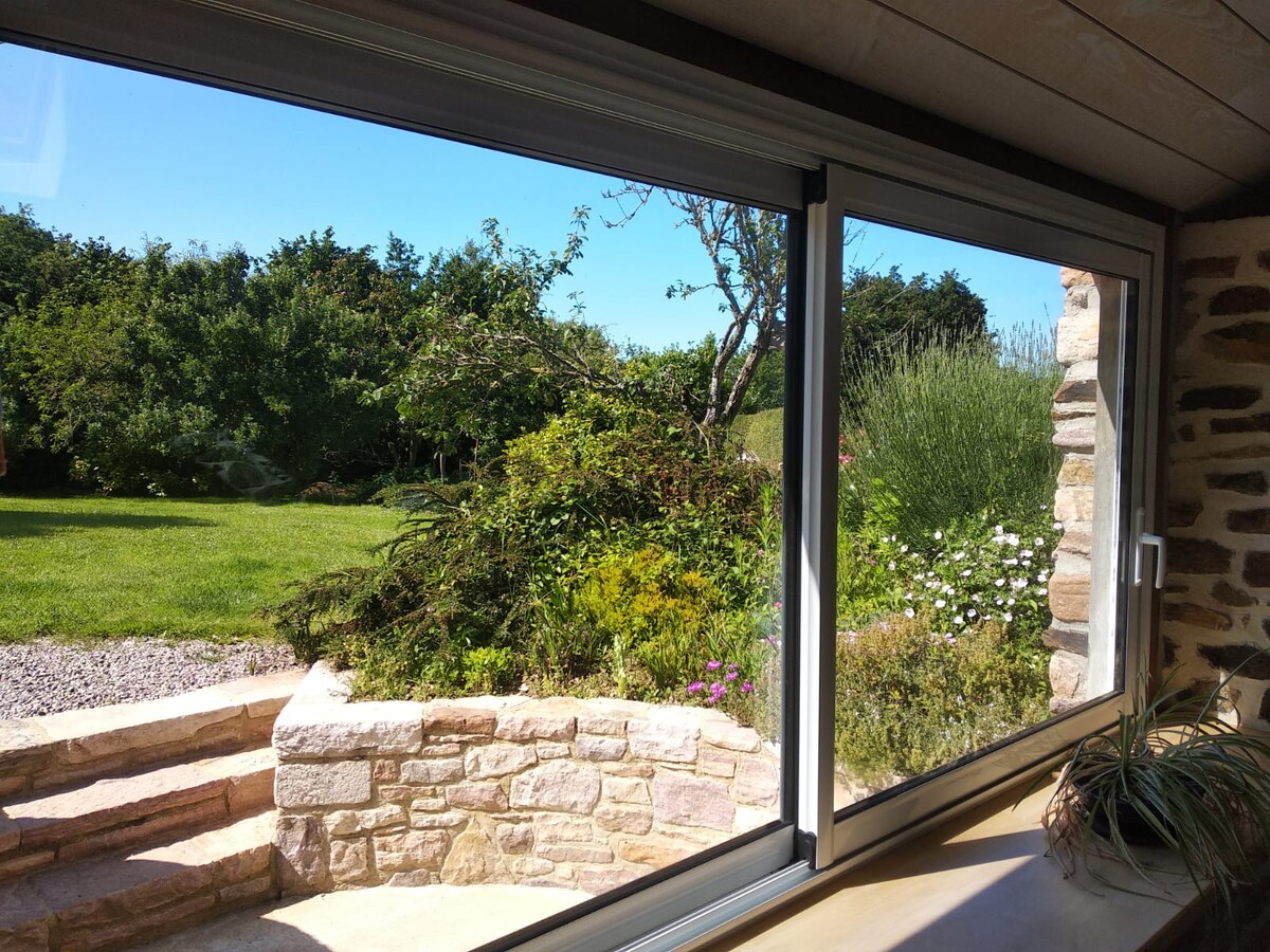 A large window offers a clear view of the lush garden outside. Vibrant greenery is seen, with flowers adding color to the landscape. The stone walls and steps in the foreground provide a natural frame for the scene, enhancing the connection between indoor and outdoor spaces.