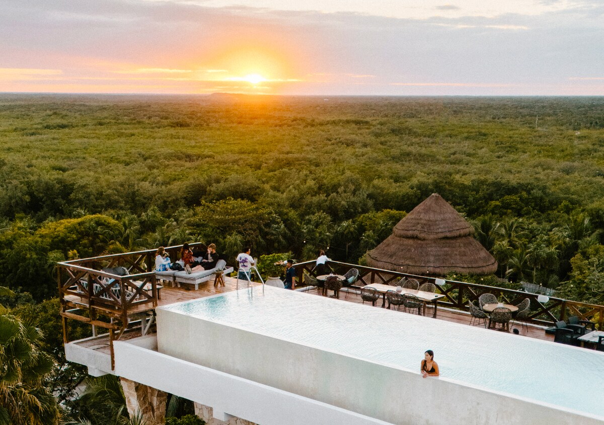 An expansive infinity pool extends towards the horizon, offering a seamless view of the sunset over lush greenery. Lounge chairs and tables are set around the pool area, where guests are seen enjoying the tranquil atmosphere. A thatched-roof structure is visible in the distance.