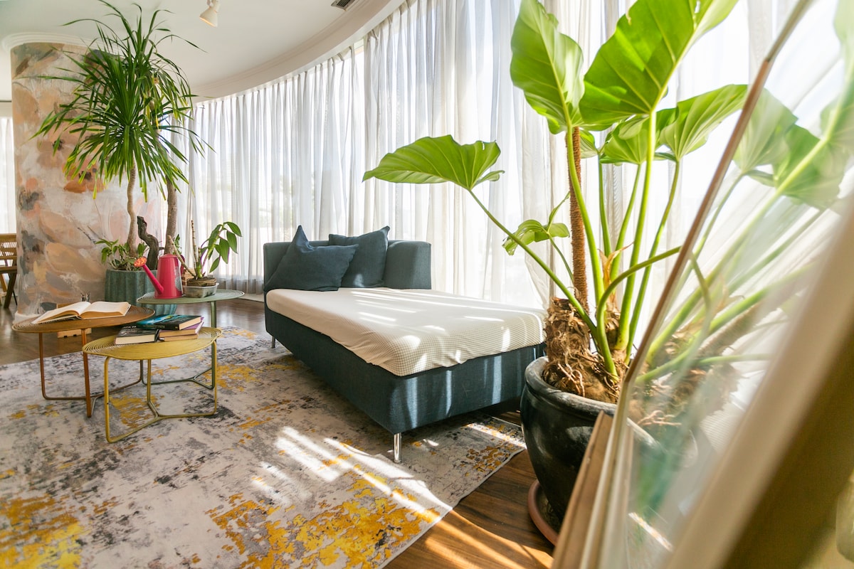 A bright seating area is highlighted by large floor-to-ceiling windows, allowing ample natural light. A modern, dark upholstered chair is paired with a low, circular table displaying books and decorative items. Lush green plants add a refreshing touch to the serene atmosphere.
