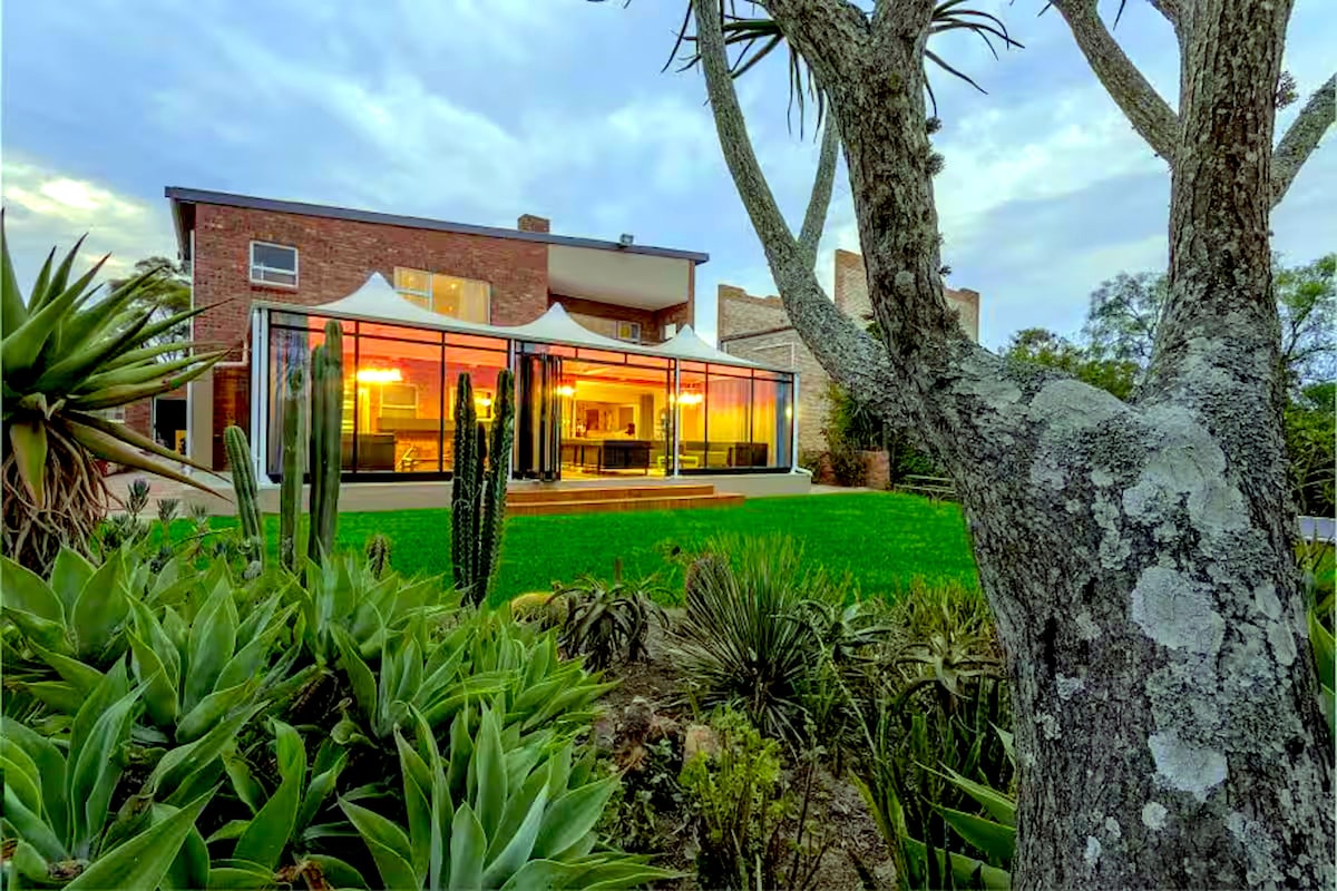 The exterior of a modern house is framed by lush greenery, featuring large glass panels that reflect warm lighting from inside. A prominent tree and various plants enhance the landscape, creating a serene outdoor space.