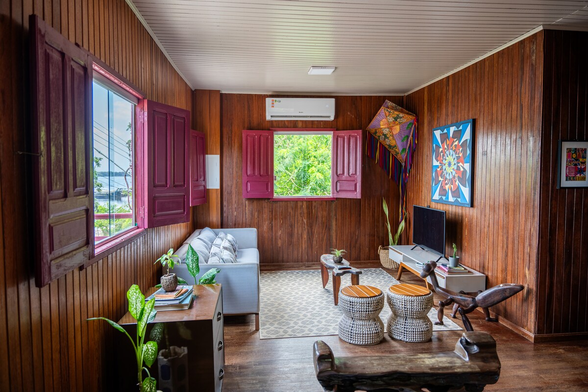 The living area features wooden walls and large windows with maroon shutters, allowing natural light to illuminate the space. A light gray sofa is positioned beside a decorative wooden table, accompanied by two unique stools. Indoor plants add a touch of greenery to the ambiance.