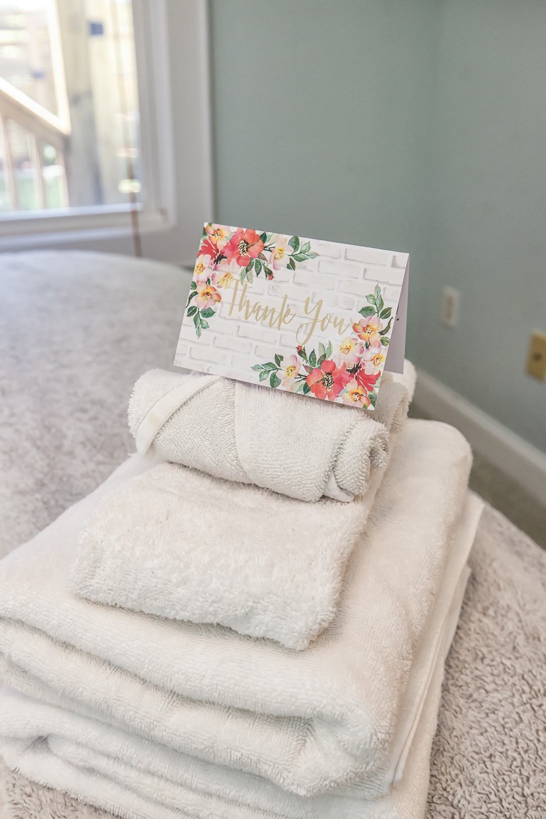 A stack of soft, white towels is displayed on a light-colored surface. A card with a floral design and the words 'Thank You' is placed on top. Natural light streams in from a nearby window, creating a serene atmosphere.