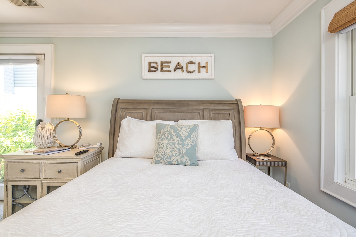 A well-appointed bedroom features a spacious bed with a white quilt and a decorative pillow. Soft light is provided by bedside lamps. A sign reading 'BEACH' adorns the wall above the bed, and large windows allow natural light to enhance the serene atmosphere.