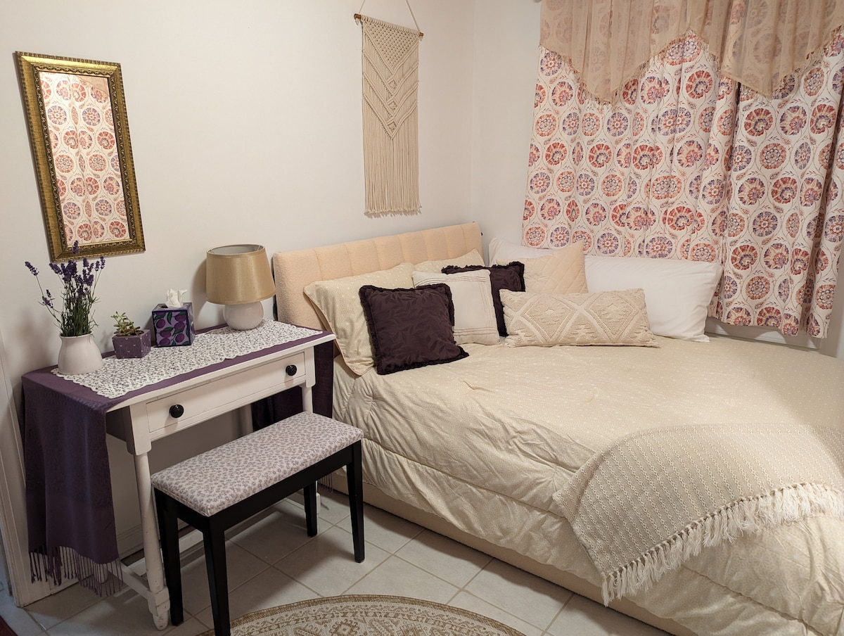 A cozy bedroom features a queen bed adorned with soft linens and decorative pillows. A small desk with a stool sits beside the bed. Natural light filters through patterned curtains, while a decorative mirror complements the warm color palette.