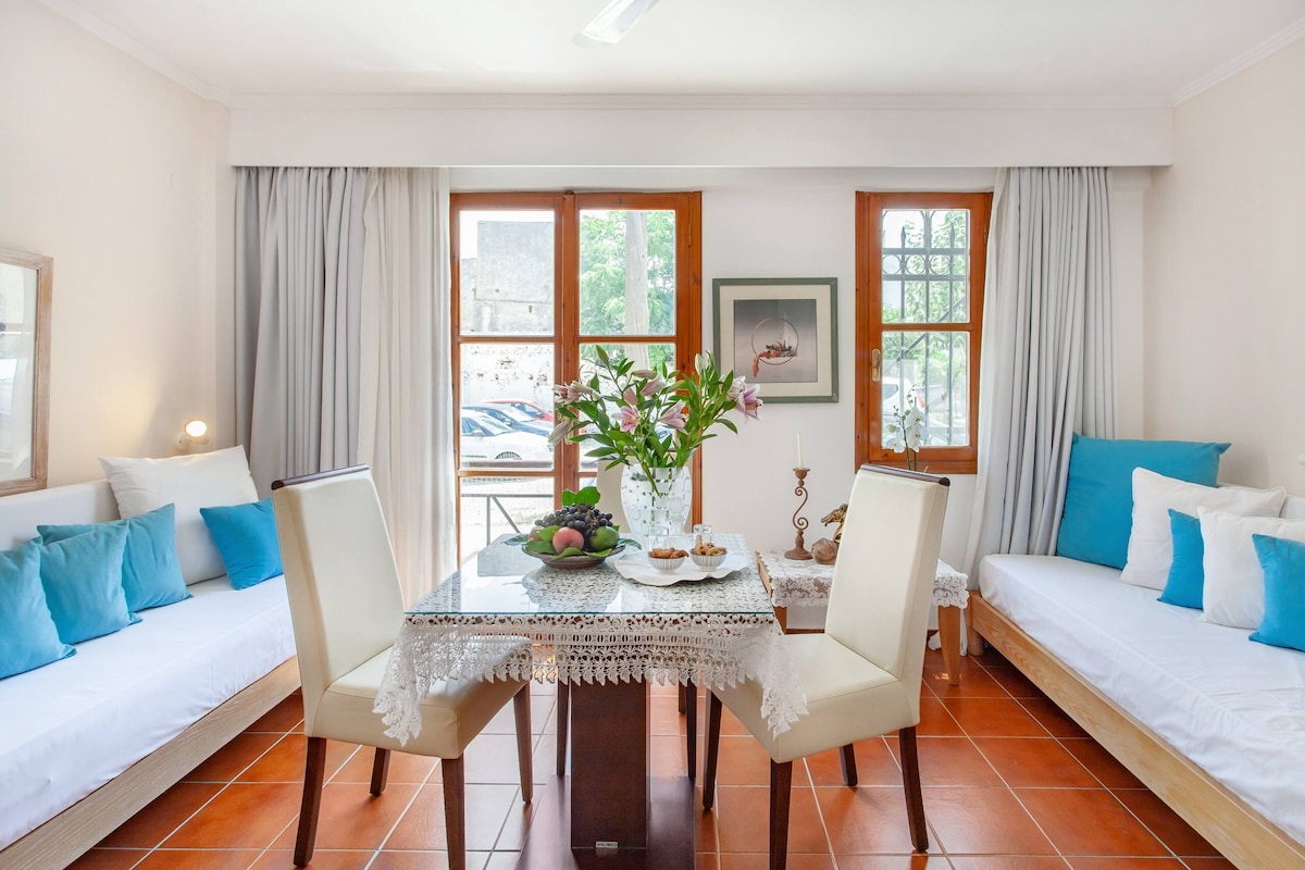 A bright seating area is furnished with two single beds and a dining table adorned with fresh flowers and a selection of fruits. Natural light streams through large windows dressed with light-colored curtains, creating a welcoming and airy environment.