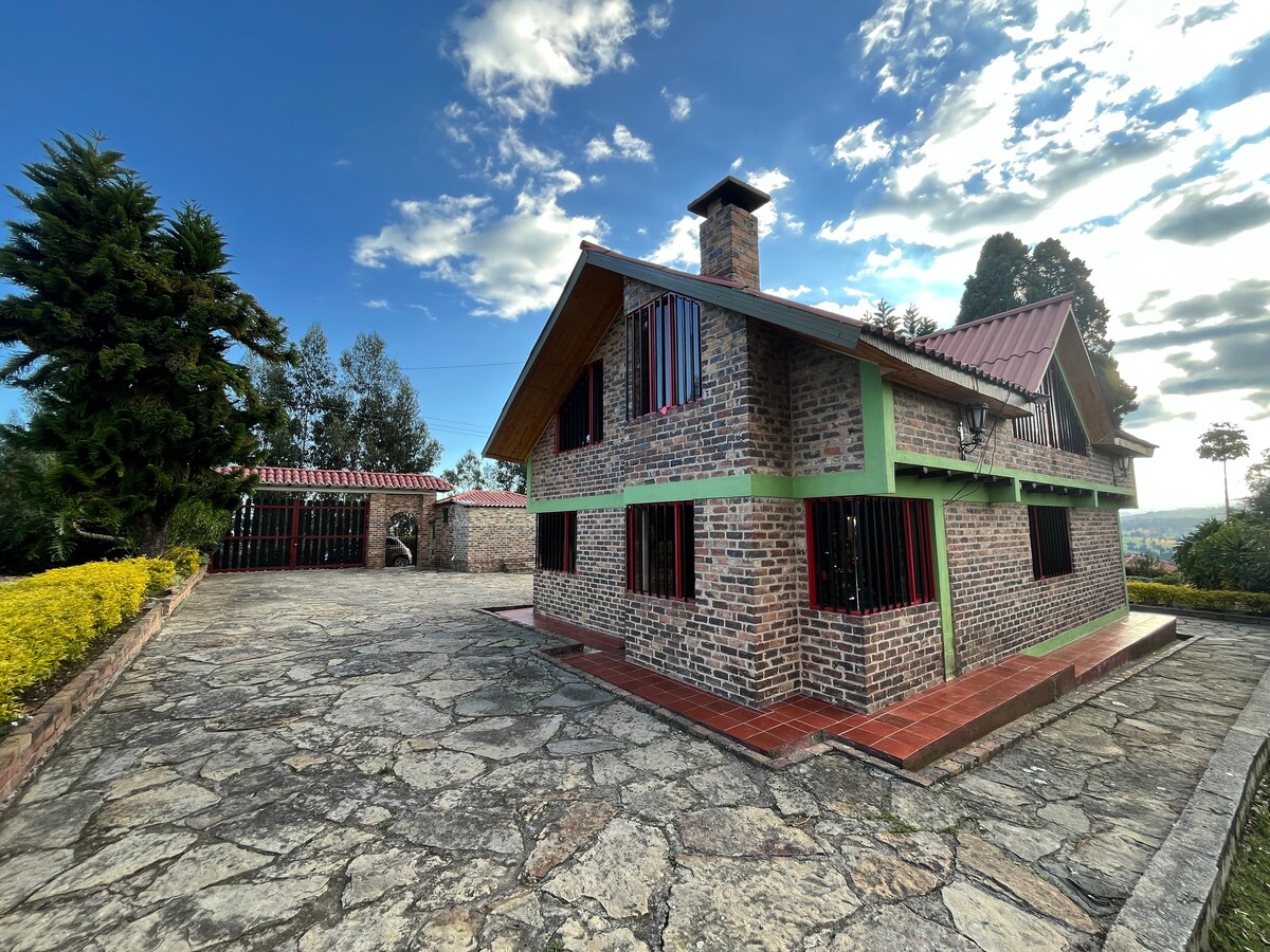 The exterior of the two-story brick house is showcased with a red-tiled roof and green trim. The stone pathway leads to the entrance. Surrounding vegetation includes tall trees and neatly trimmed hedges. A view of the nearby landscape is visible in the background.