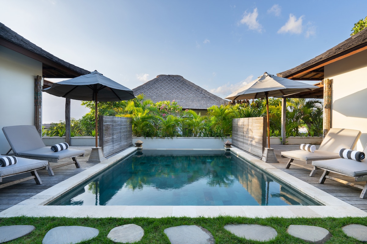 A serene pool area is surrounded by lush greenery and sun loungers. Two umbrellas provide shade over the loungers positioned beside the calm water. The reflection of the sky is visible on the pool's surface, enhancing the tranquil ambiance of the space.