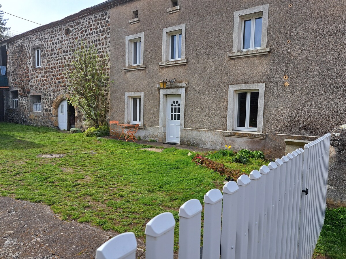 The renovated farmhouse exterior features a façade of stone and neutral-colored walls. Windows with white frames are positioned symmetrically, enhancing the building's character. A small garden area with grass is visible, complemented by a white picket fence that encloses the property.