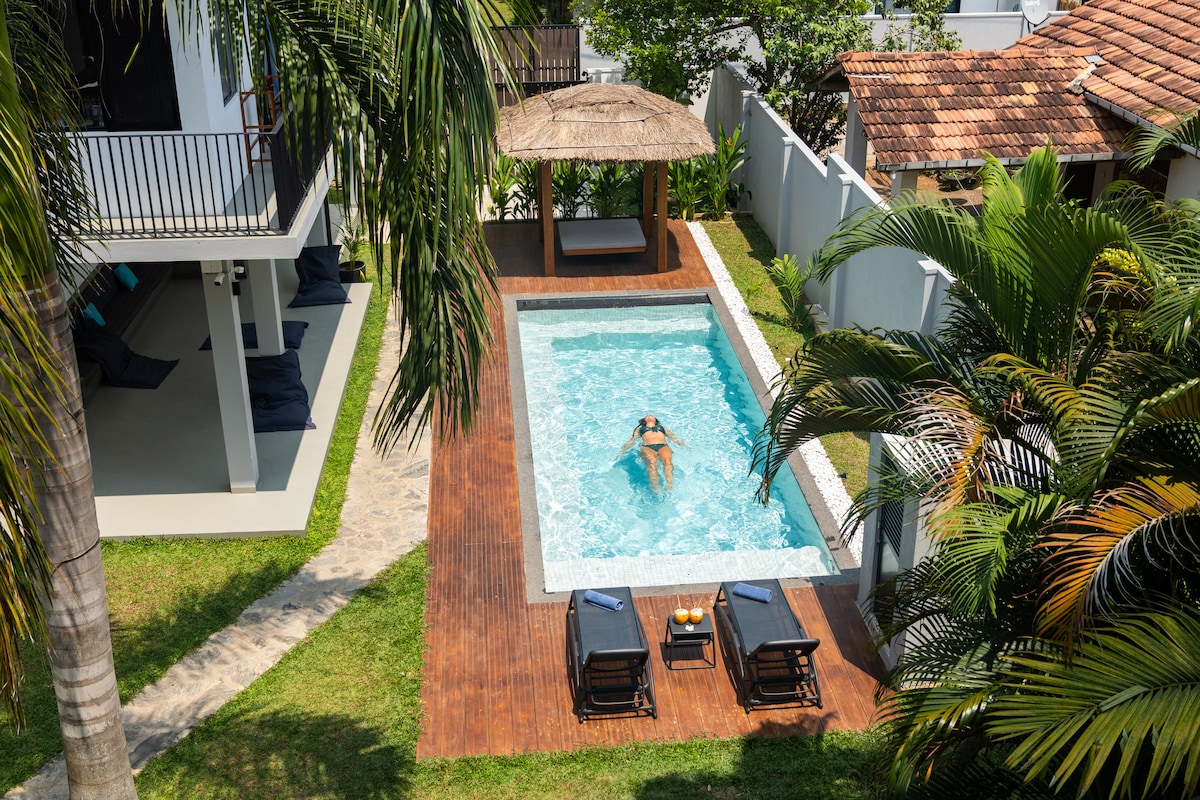 A private pool area is surrounded by lush tropical greenery. Sun loungers are positioned alongside the sparkling water, with a thatched-roof gazebo providing shade. The space offers a serene environment for relaxation and outdoor enjoyment.