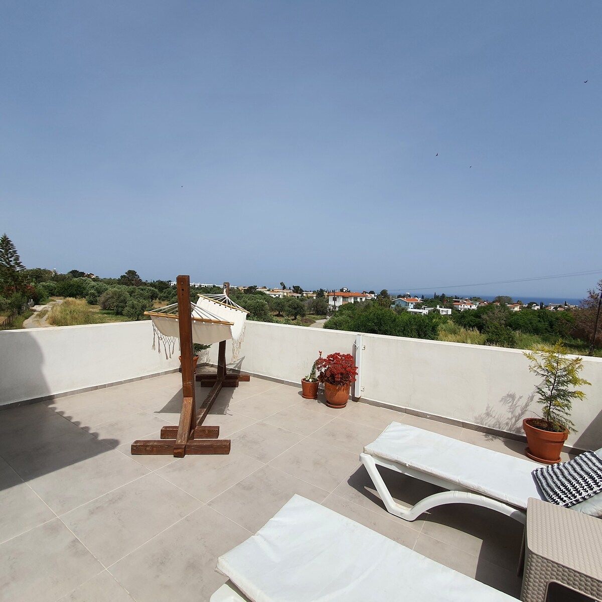 An outdoor terrace features lounge chairs and a hanging chair on a tiled surface. Potted plants add greenery to the space, with a distant view of the mountains and sea visible beyond the white boundary wall.