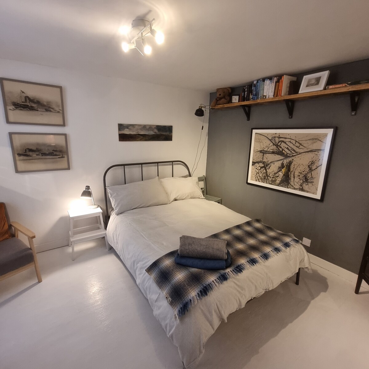 A cozy bedroom features a spacious bed with a folded blanket positioned at the foot. Neutral walls are accented by framed artwork and decorative shelving. A bedside lamp and a small armchair add functional elements, while natural light illuminates the space.