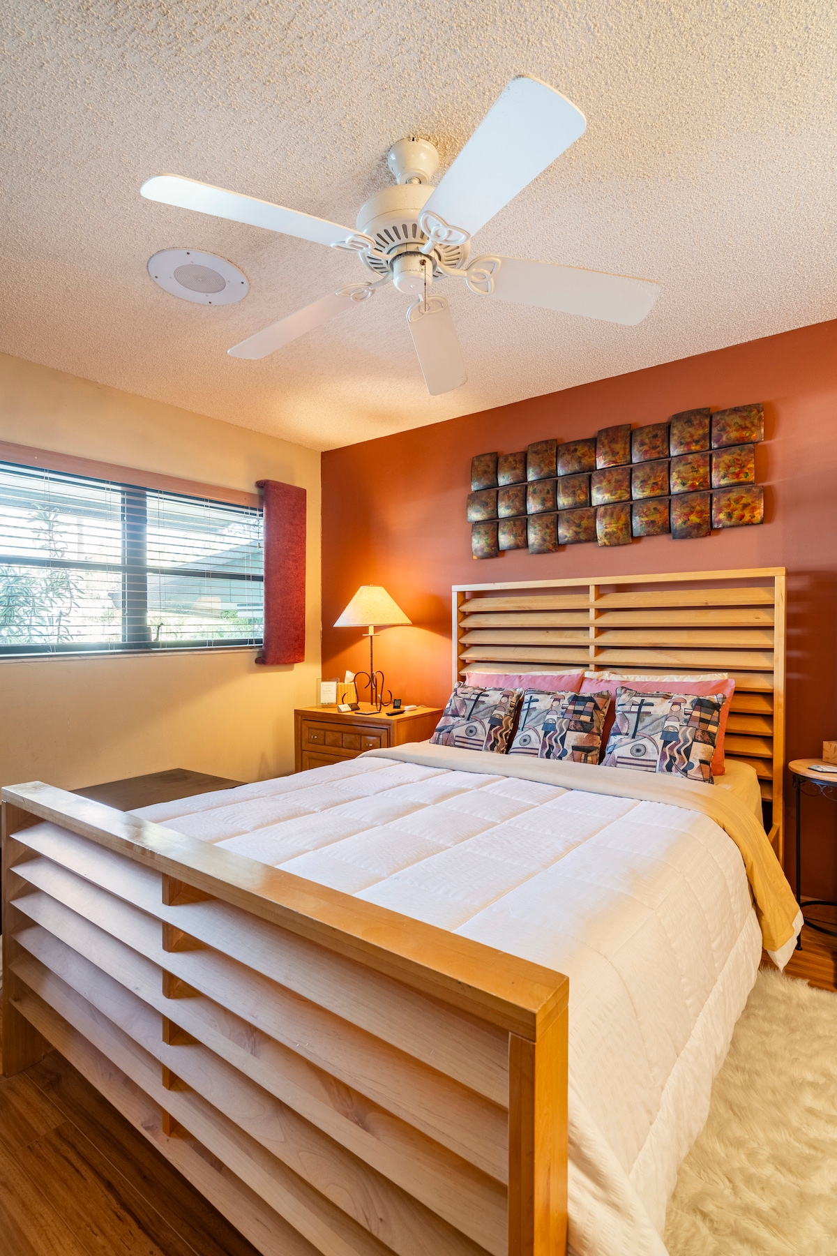 A spacious bedroom is captured with a wooden bed frame featuring a modern design. The bed is dressed in a white quilt, and decorative pillows add a touch of color. A bedside table holds a lamp, while natural light filters through a window with blinds.