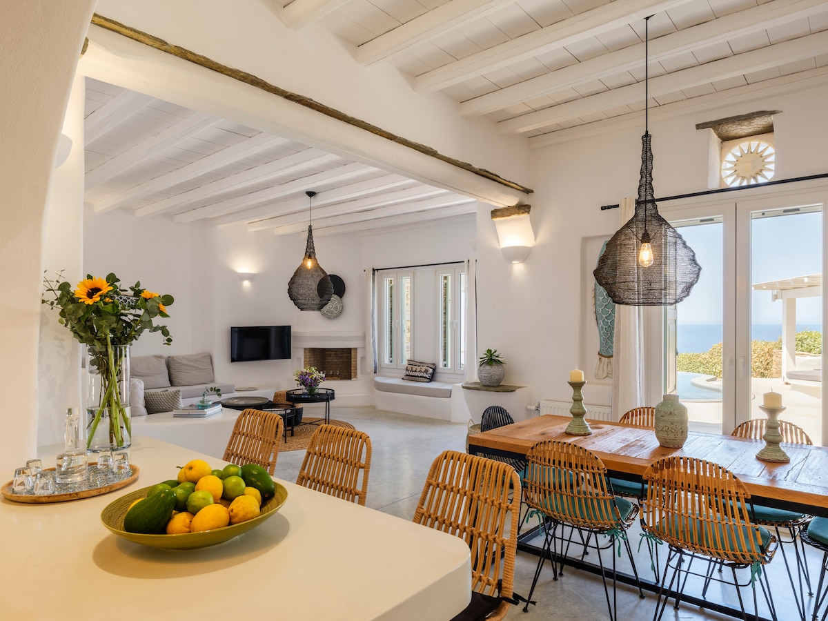 A spacious living area features a large dining table surrounded by stylish woven chairs. Natural light floods in through glass doors, offering views of the surrounding landscape. Decorative light fixtures hang from the ceiling, and a cozy seating area with a fireplace is visible in the background.