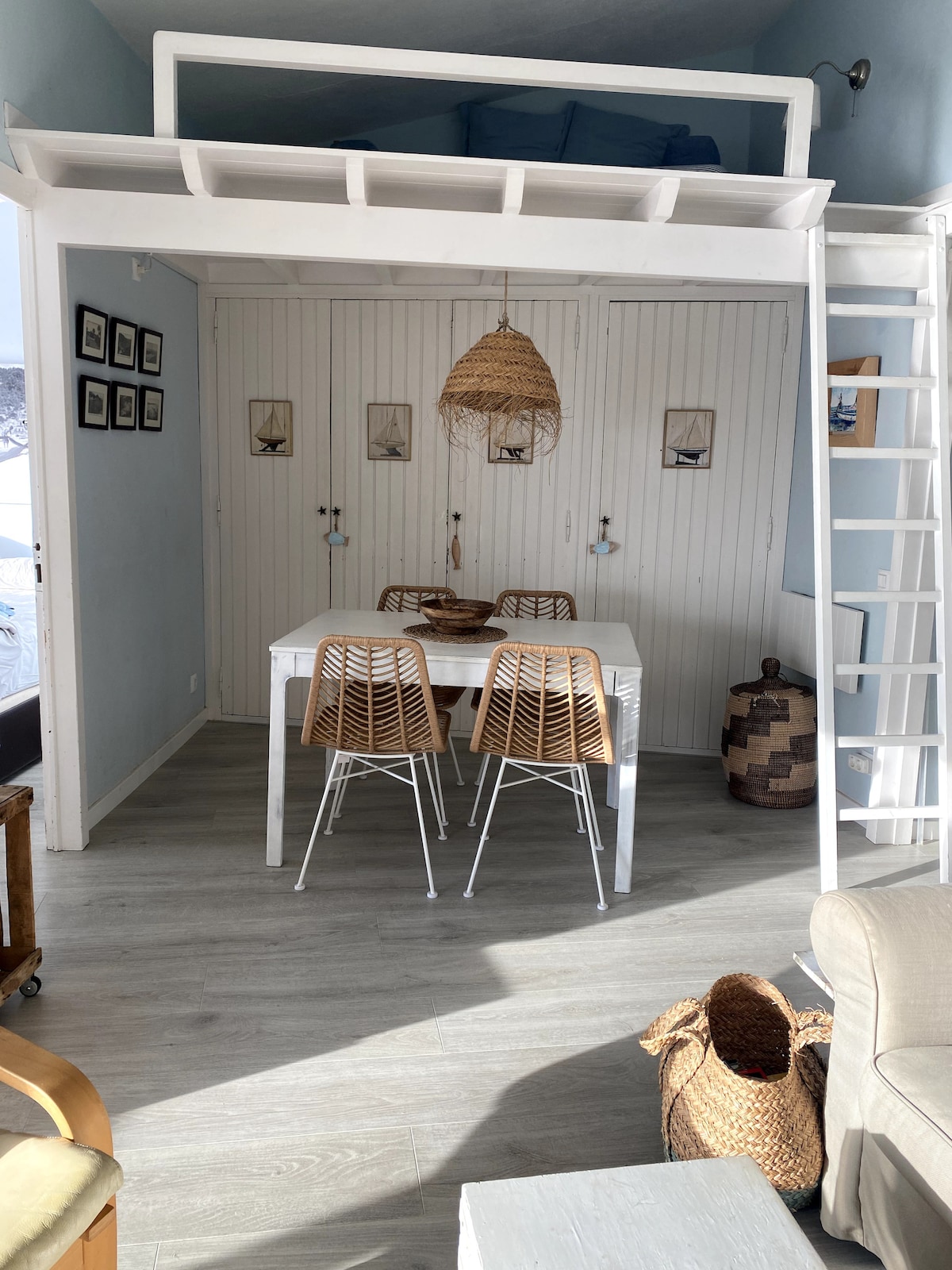 A bright dining area features a white table surrounded by four woven chairs. A ladder leads to a lofted sleeping area above, while natural light pours in through a large window adjacent to the seating area, showcasing light blue walls and accented with neutral decor.